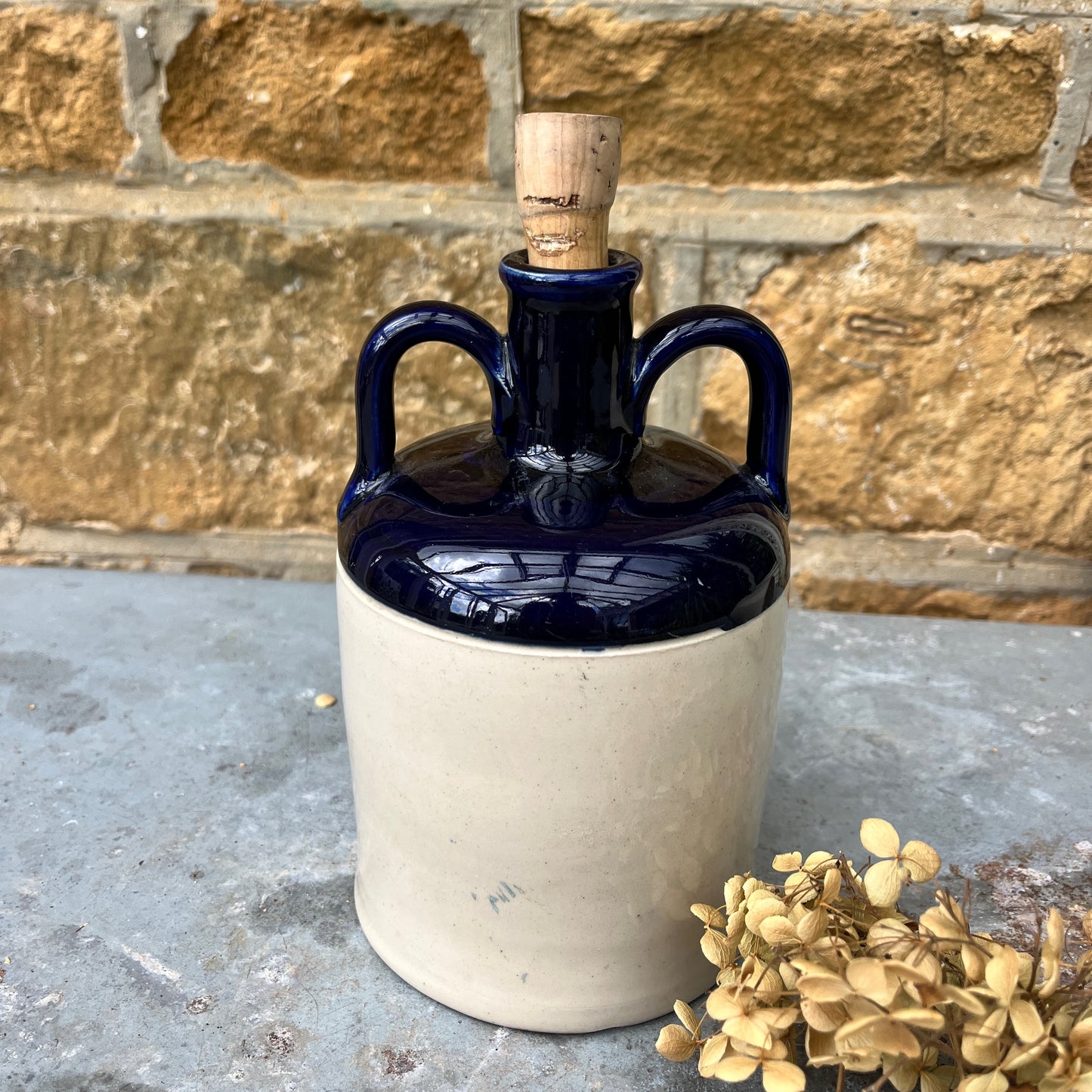 A stoneware flagon with a robust, earthy brown base transitioning to a glossy blue glaze on the top half. The flagon features two sturdy handles and a narrow stoppered spout, exuding rustic charm