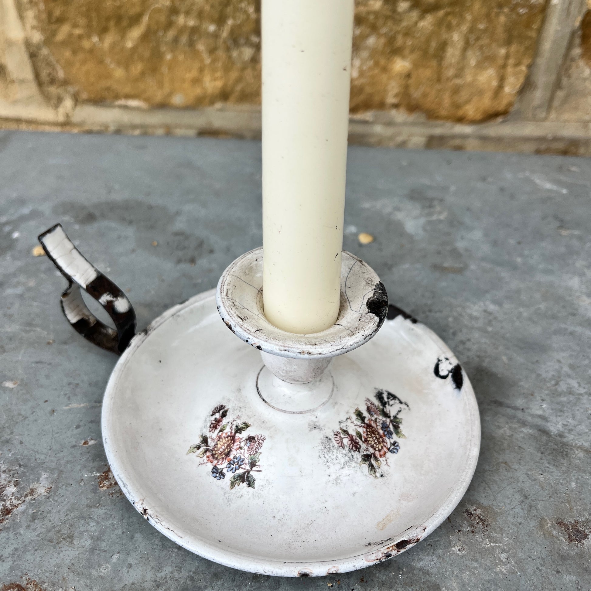 A white enamel vintage French candle holder with a decorative flowers and muted finish, featuring a shallow, round dish to hold a tapered candle. The holder has a smooth, curved edge, exuding classic French charm