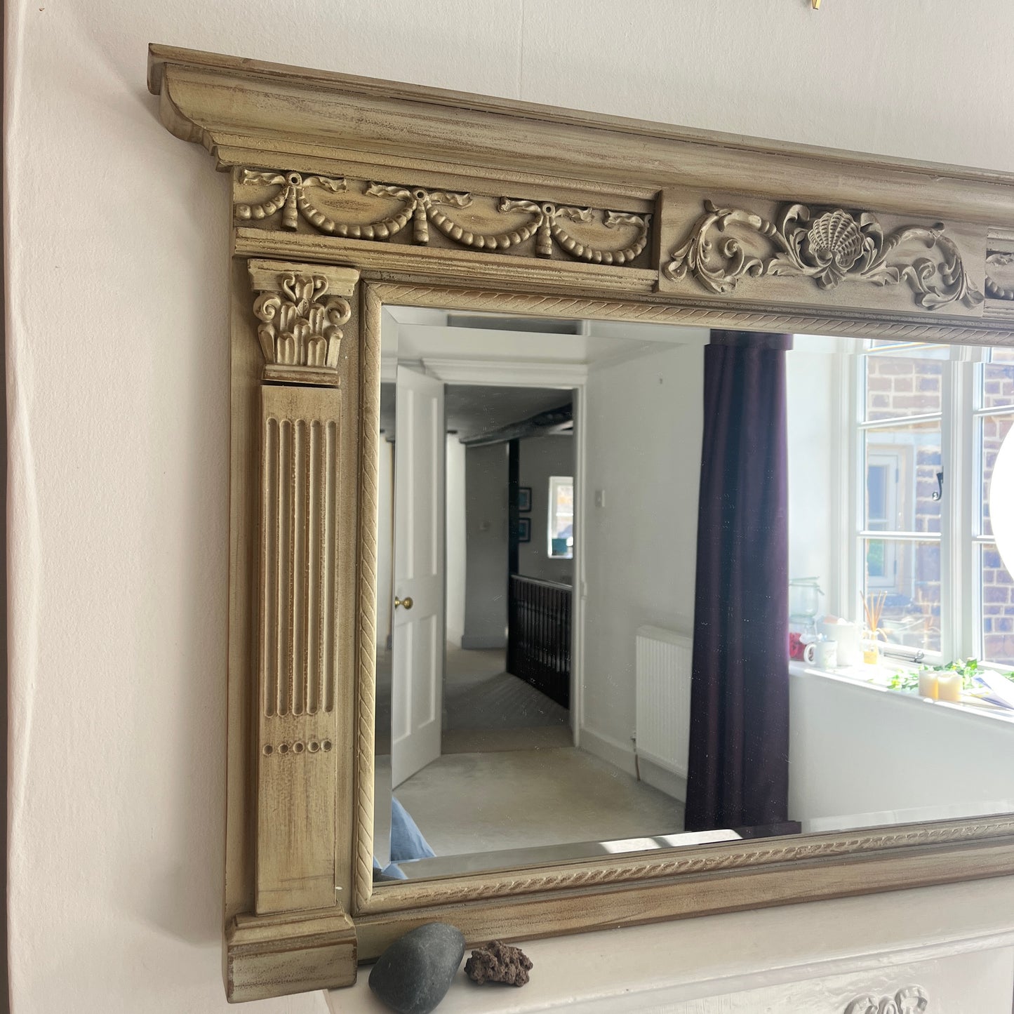 A classic over mantle mirror featuring a grand wooden frame with Romanesque-style columns on either side, adorned with intricate carvings. The beveled mirror surface adds depth and elegance, with a distressed cream finish, exuding luxurious vintage charm