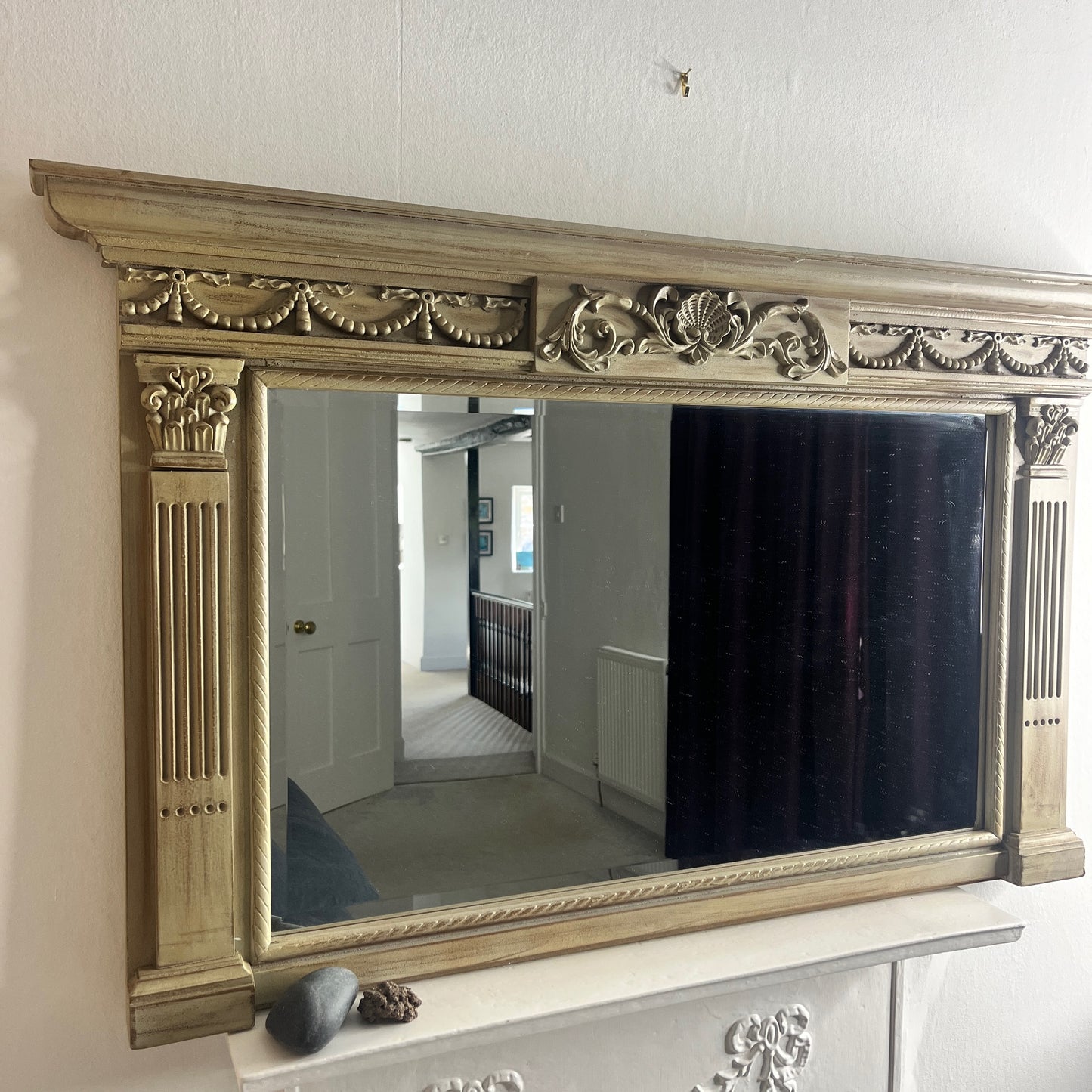 A classic over mantle mirror featuring a grand wooden frame with Romanesque-style columns on either side, adorned with intricate carvings. The beveled mirror surface adds depth and elegance, with a distressed cream finish, exuding luxurious vintage charm