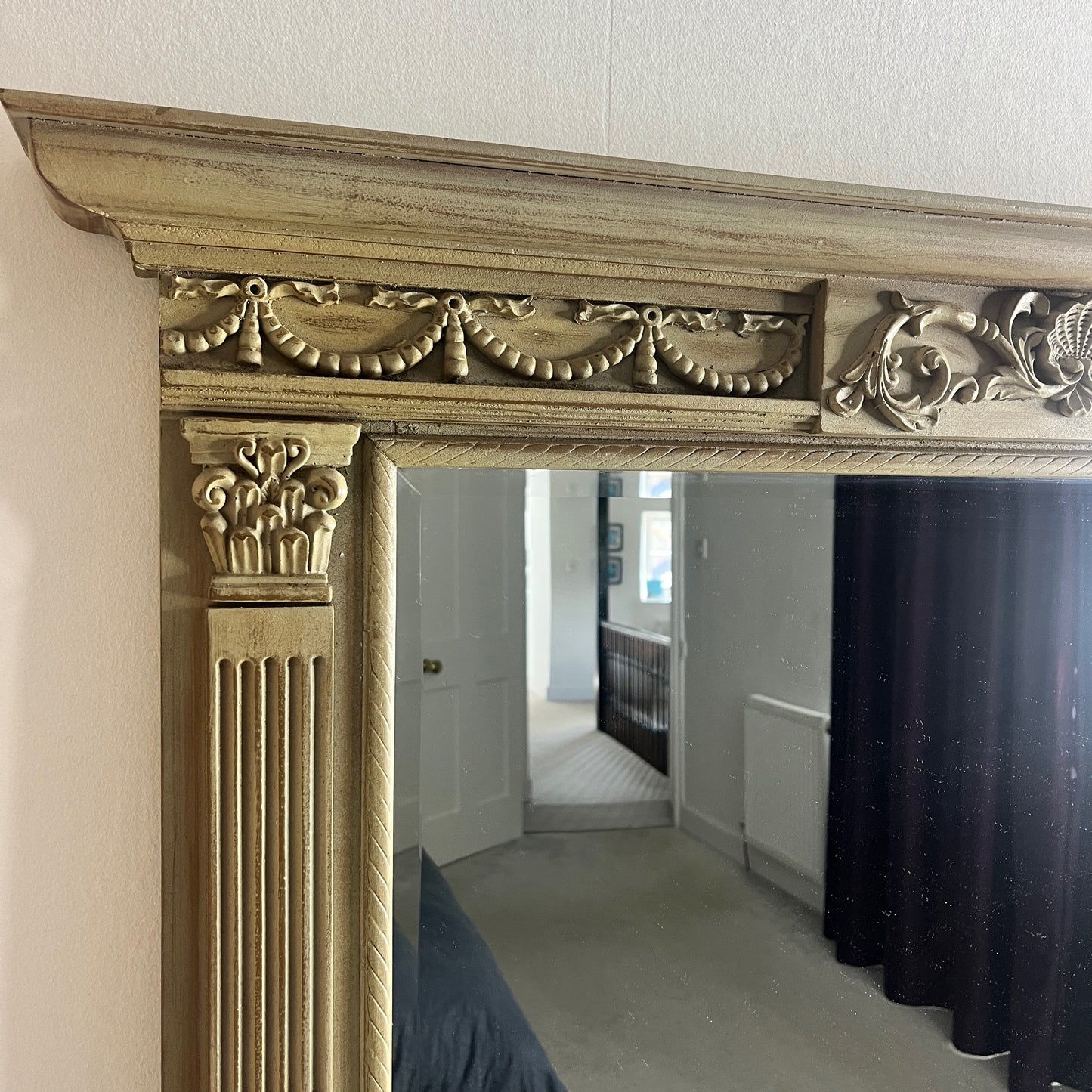 A classic over mantle mirror featuring a grand wooden frame with Romanesque-style columns on either side, adorned with intricate carvings. The beveled mirror surface adds depth and elegance, with a distressed cream finish, exuding luxurious vintage charm