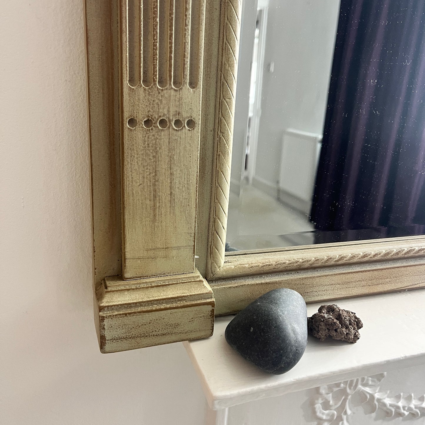 A classic over mantle mirror featuring a grand wooden frame with Romanesque-style columns on either side, adorned with intricate carvings. The beveled mirror surface adds depth and elegance, with a distressed cream finish, exuding luxurious vintage charm