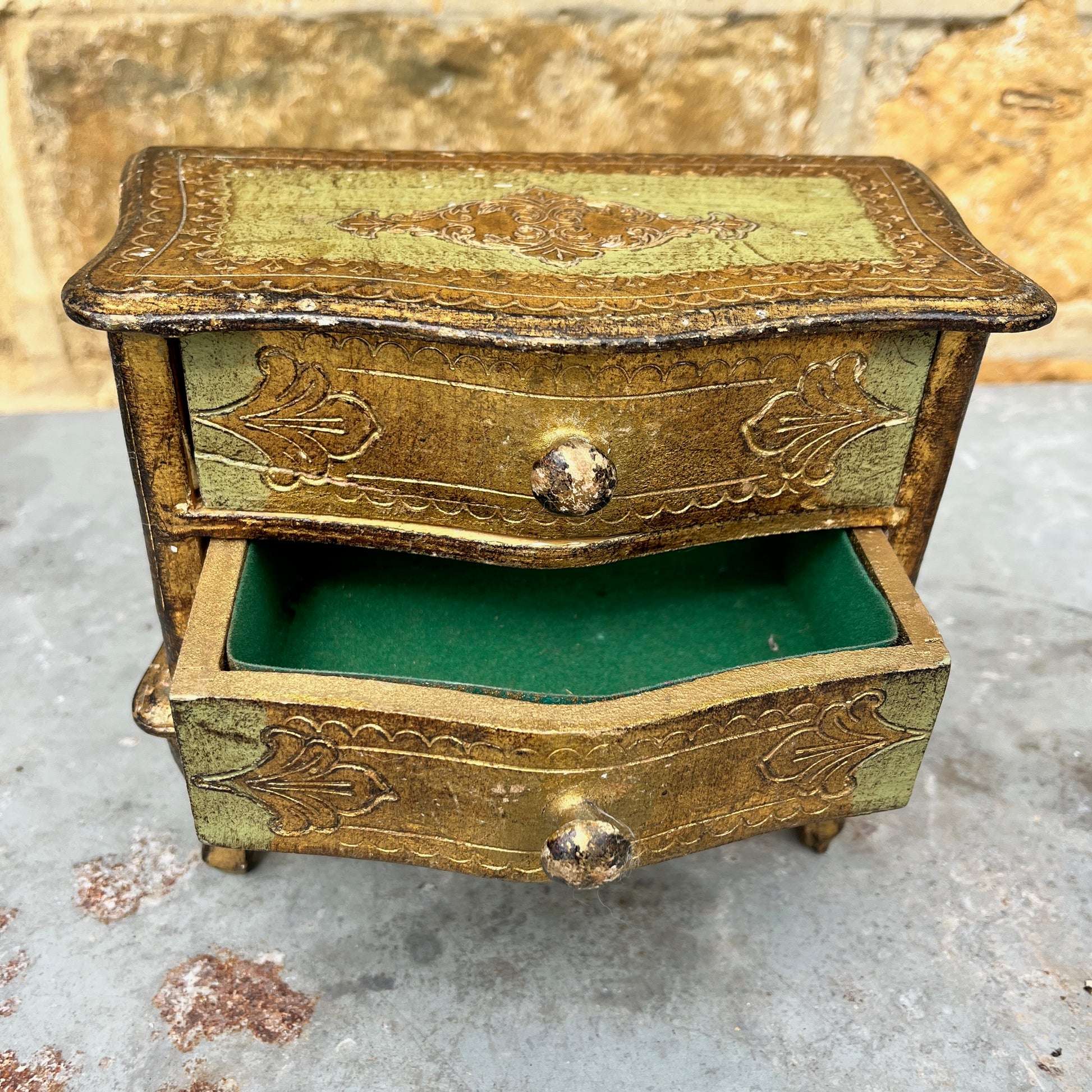 A miniature Florentine chest of drawers featuring a luxurious green and gold colour scheme. The chest has a rectangular shape with a smooth, glossy gold finish on the body, accented by green detailing along the edges. The front displays 2 small drawers, each adorned with gold handles,