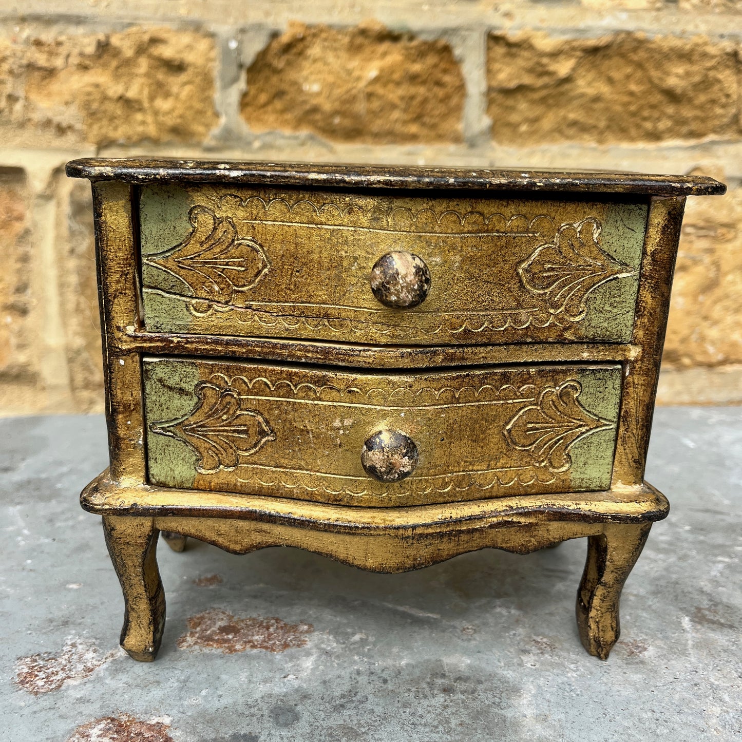 A miniature Florentine chest of drawers featuring a luxurious green and gold colour scheme. The chest has a rectangular shape with a smooth, glossy gold finish on the body, accented by green detailing along the edges. The front displays 2 small drawers, each adorned with gold handles,