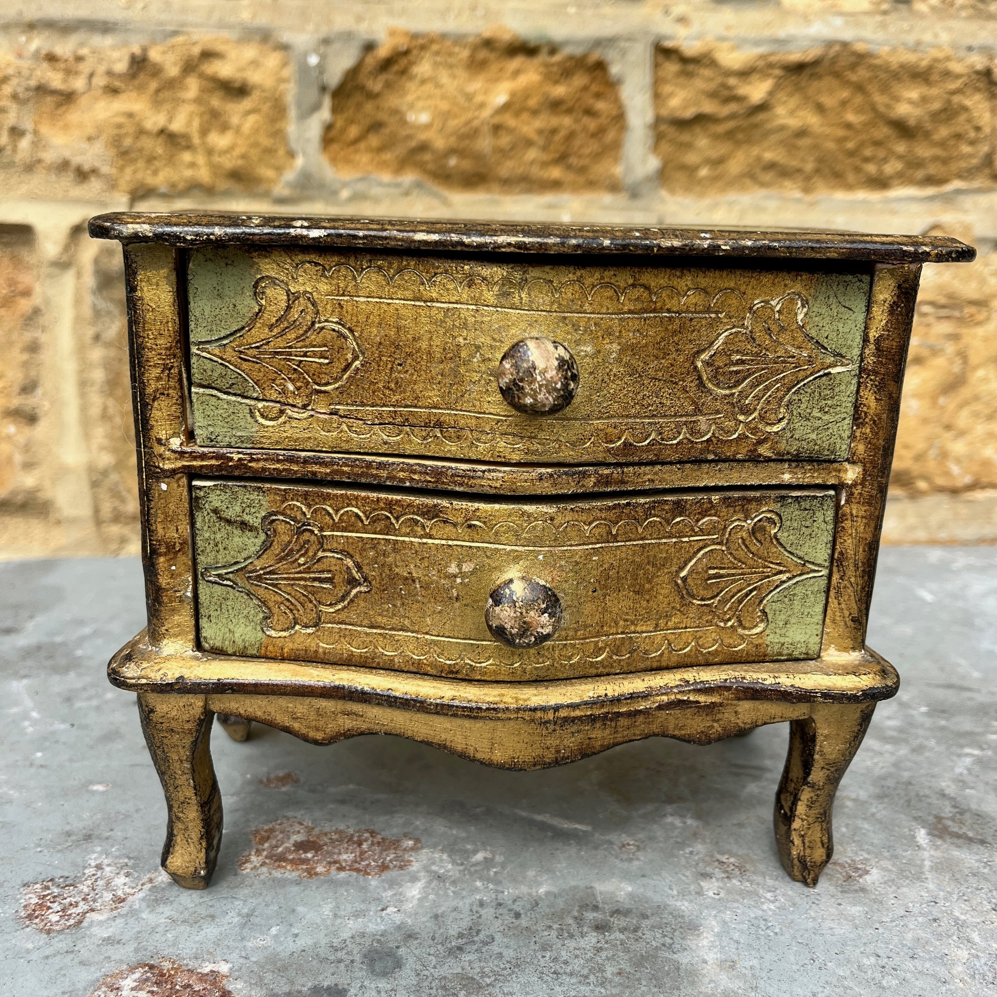 A miniature Florentine chest of drawers featuring a luxurious green and gold colour scheme. The chest has a rectangular shape with a smooth, glossy gold finish on the body, accented by green detailing along the edges. The front displays 2 small drawers, each adorned with gold handles,