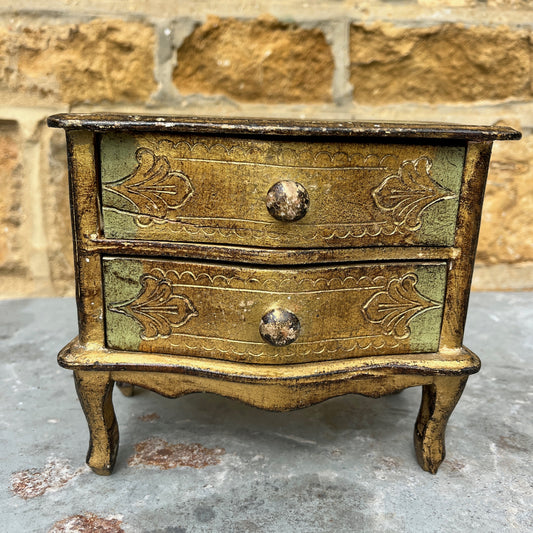 A miniature Florentine chest of drawers featuring a luxurious green and gold colour scheme. The chest has a rectangular shape with a smooth, glossy gold finish on the body, accented by green detailing along the edges. The front displays 2 small drawers, each adorned with gold handles,