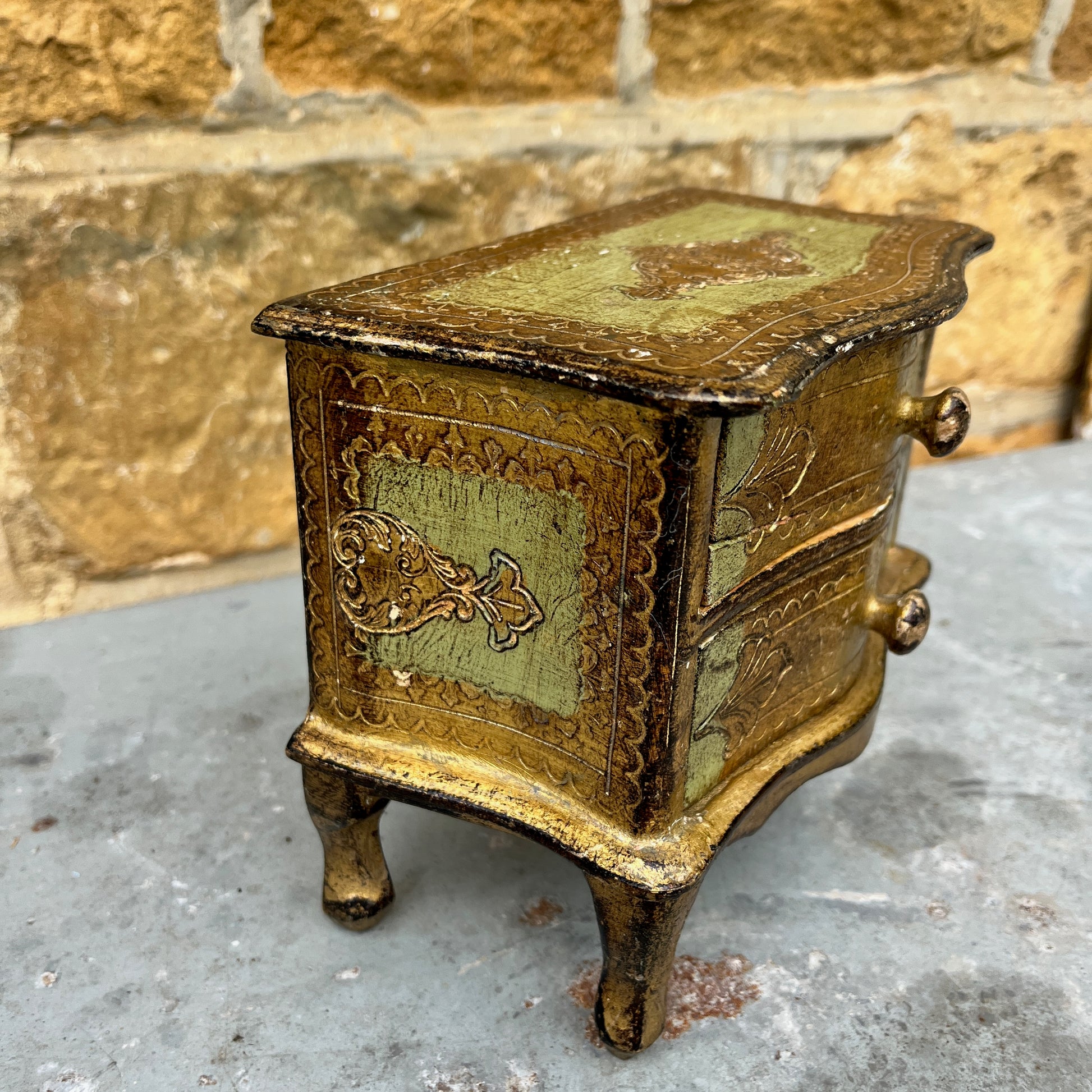 A miniature Florentine chest of drawers featuring a luxurious green and gold colour scheme. The chest has a rectangular shape with a smooth, glossy gold finish on the body, accented by green detailing along the edges. The front displays 2 small drawers, each adorned with gold handles,