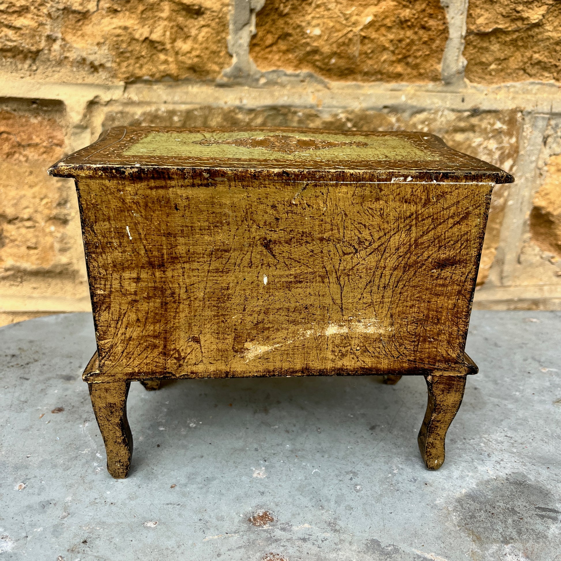 A miniature Florentine chest of drawers featuring a luxurious green and gold colour scheme. The chest has a rectangular shape with a smooth, glossy gold finish on the body, accented by green detailing along the edges. The front displays 2 small drawers, each adorned with gold handles,