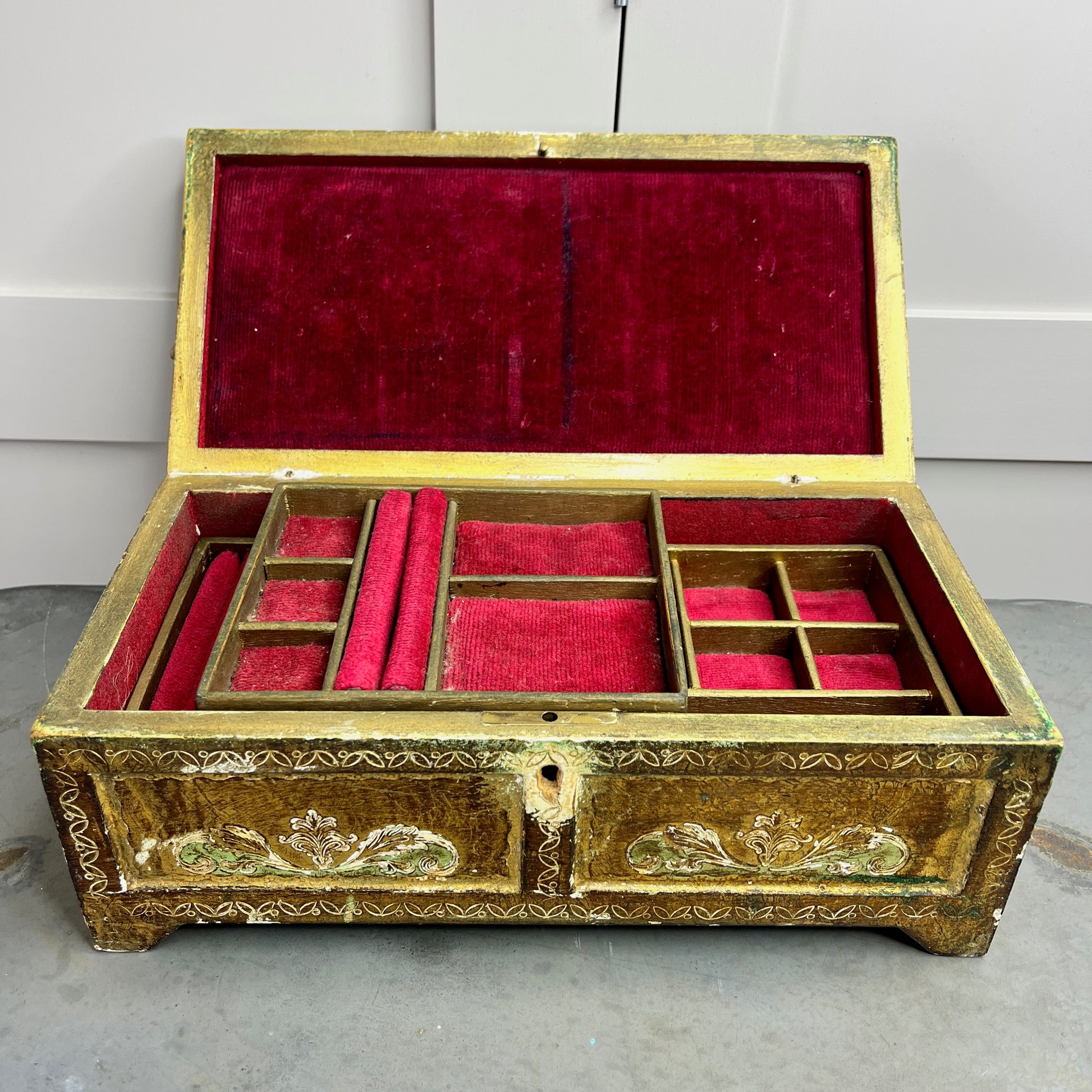 A vintage Florentine jewellery box with a rectangular shape, featuring a rich gold exterior with intricate green accents. The box is adorned with ornate, baroque-style gold detailing along the edges and corners, showcasing classic Florentine craftsmanship. The lid opens to reveal a luxurious red fabric lining.   The box stands on small gold coloured feet, exuding antique charm and sophistication.