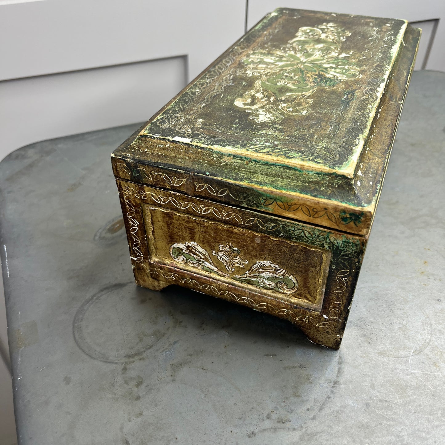A vintage Florentine jewellery box with a rectangular shape, featuring a rich gold exterior with intricate green accents. The box is adorned with ornate, baroque-style gold detailing along the edges and corners, showcasing classic Florentine craftsmanship. The lid opens to reveal a luxurious red fabric lining.   The box stands on small gold coloured feet, exuding antique charm and sophistication.
