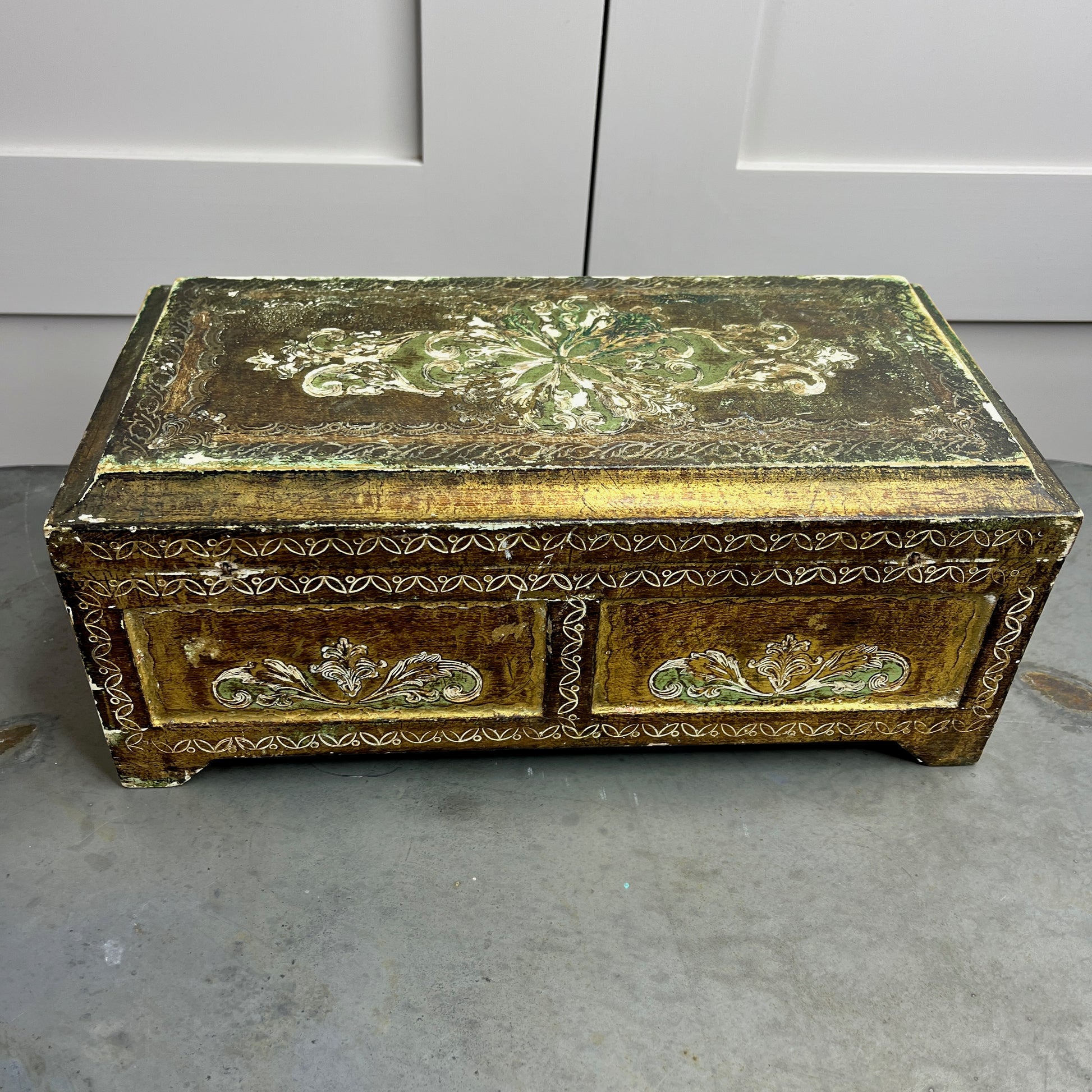 A vintage Florentine jewellery box with a rectangular shape, featuring a rich gold exterior with intricate green accents. The box is adorned with ornate, baroque-style gold detailing along the edges and corners, showcasing classic Florentine craftsmanship. The lid opens to reveal a luxurious red fabric lining.   The box stands on small gold coloured feet, exuding antique charm and sophistication.