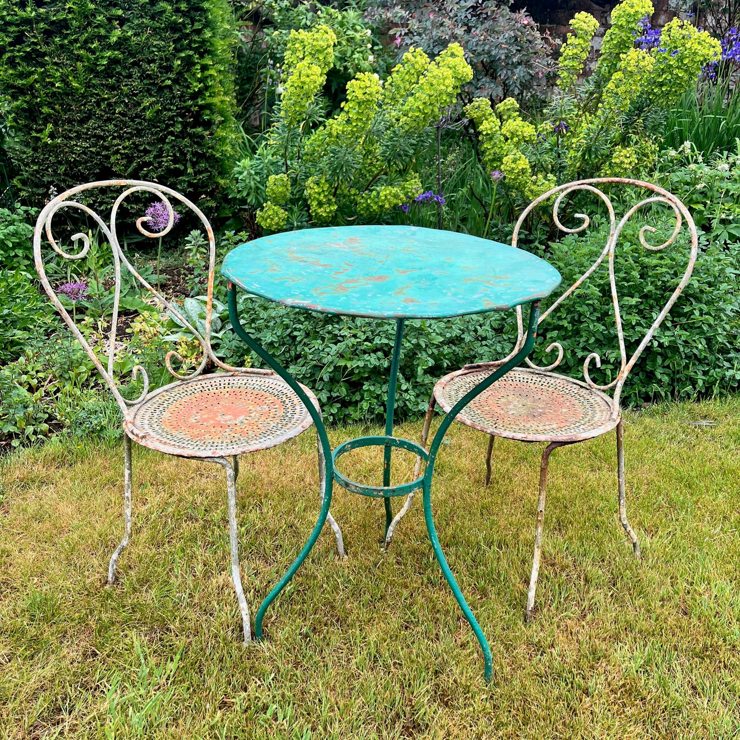 A French vintage bistro set featuring a round table with a green and orange weathered finish, paired with matching chairs that show a distressed, rustic texture. The set exudes classic outdoor charm, with the table’s smooth surface and the chairs’ slightly worn frames