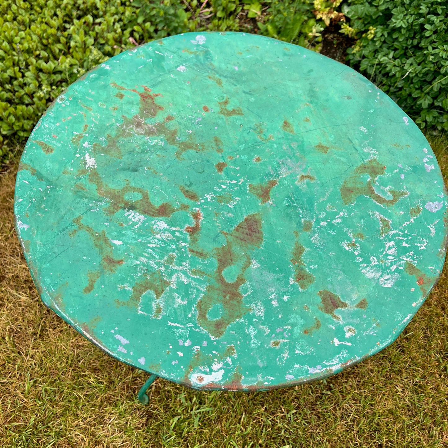 A French vintage bistro set featuring a round table with a green and orange weathered finish, paired with matching chairs that show a distressed, rustic texture. The set exudes classic outdoor charm, with the table’s smooth surface and the chairs’ slightly worn frames