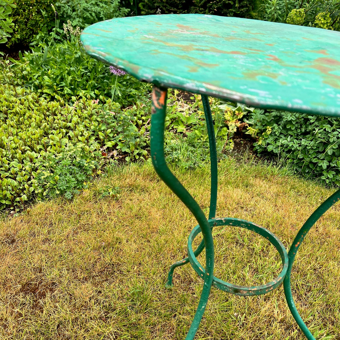 A French vintage bistro set featuring a round table with a green and orange weathered finish, paired with matching chairs that show a distressed, rustic texture. The set exudes classic outdoor charm, with the table’s smooth surface and the chairs’ slightly worn frames