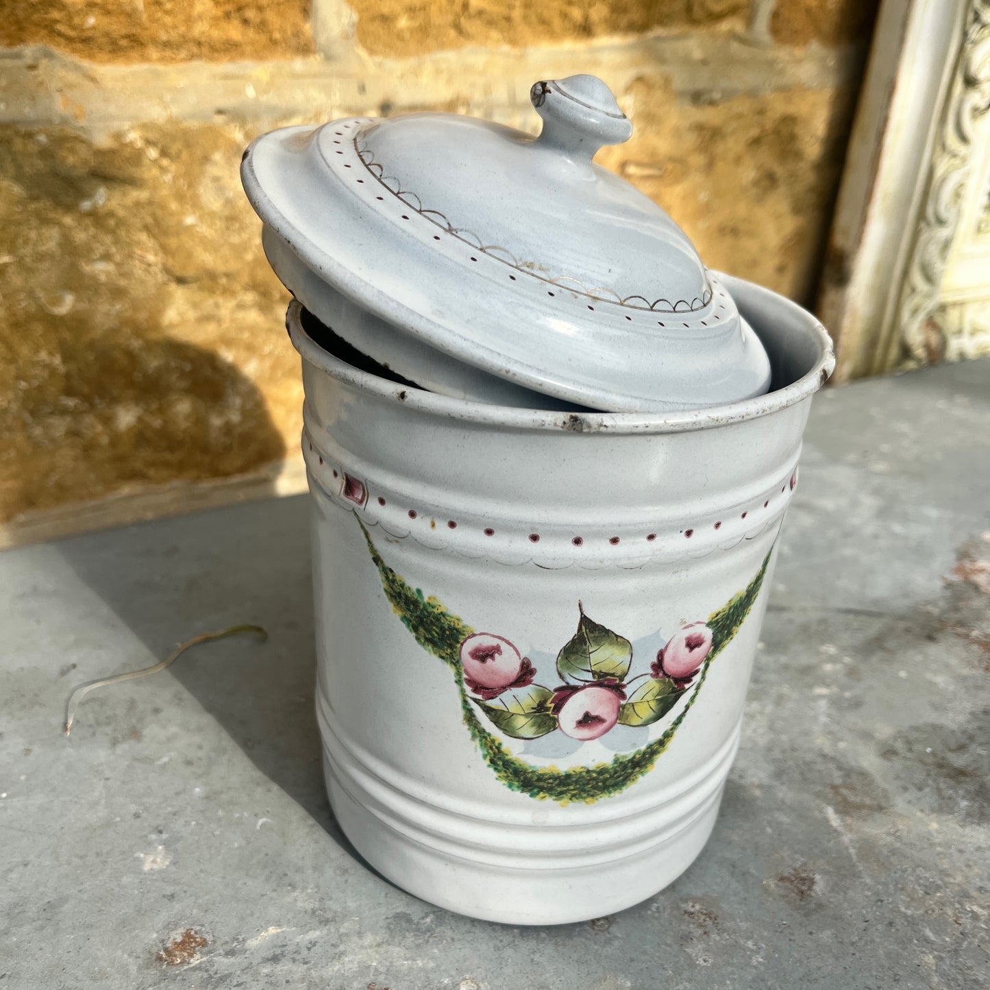 A vintage French enamel canister with a glossy white finish, adorned with intricate gold detailing and a colorful floral pattern on the front.