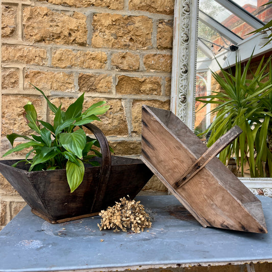 A vintage wooden French trug, featuring a shallow, rectangular shape with a sturdy central handle. The trug has a weathered, natural finish,  exuding classic gardening charm