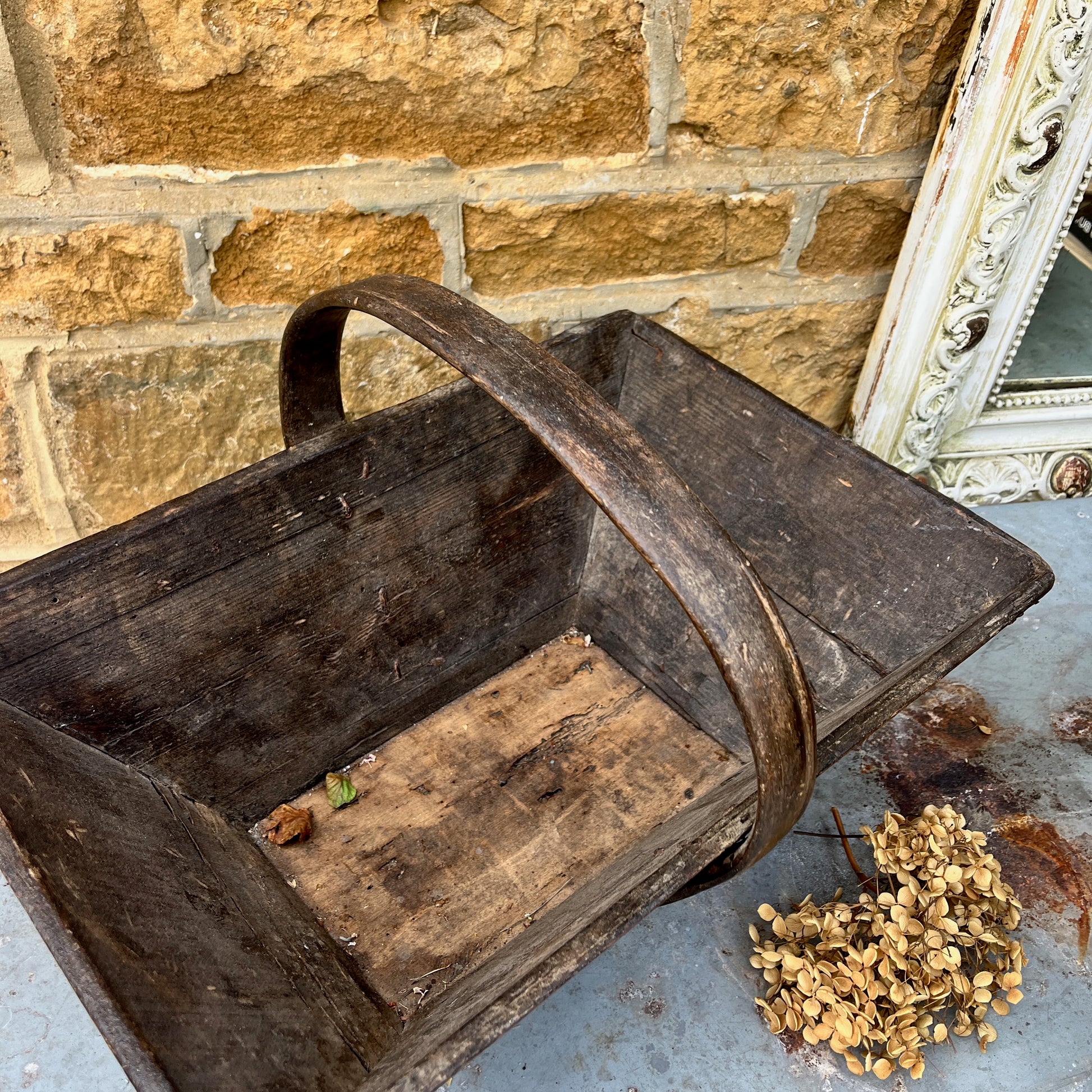A vintage wooden French trug, featuring a shallow, rectangular shape with a sturdy central handle. The trug has a weathered, natural finish,  exuding classic gardening charm