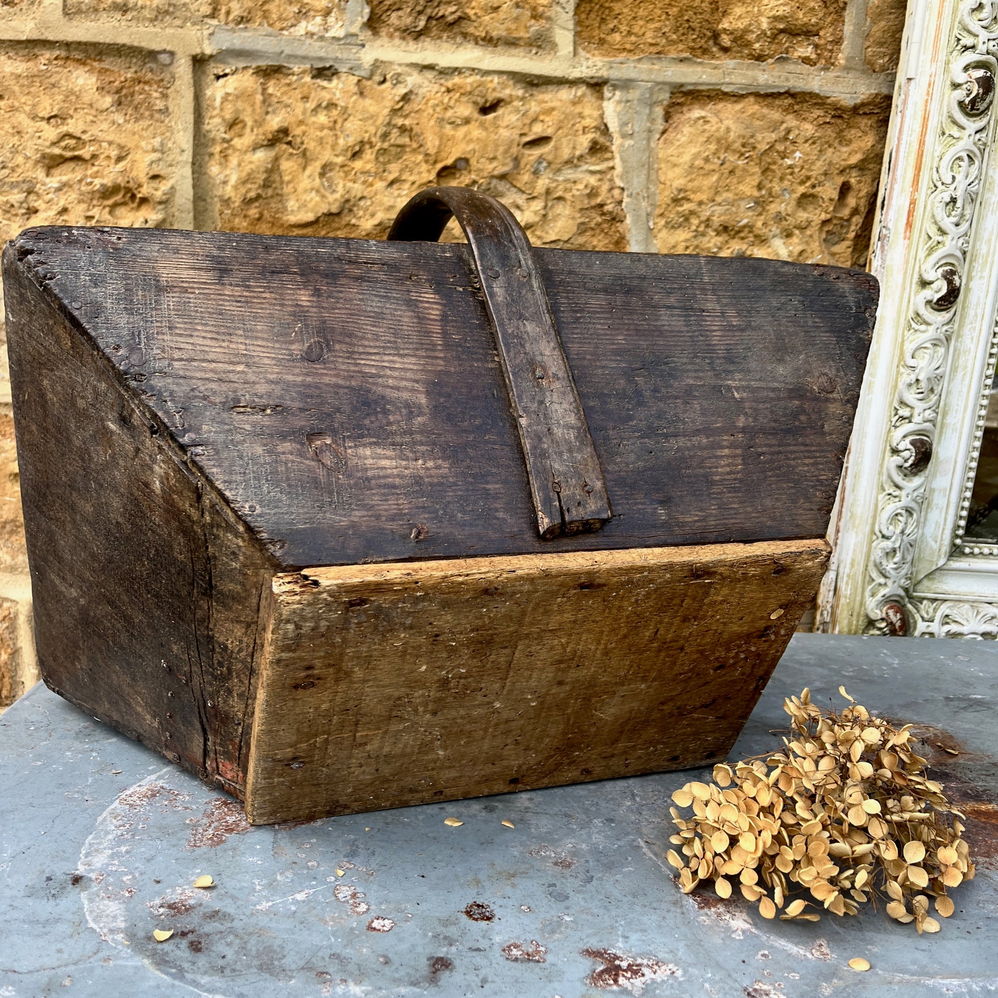 A vintage wooden French trug, featuring a shallow, rectangular shape with a sturdy central handle. The trug has a weathered, natural finish,  exuding classic gardening charm