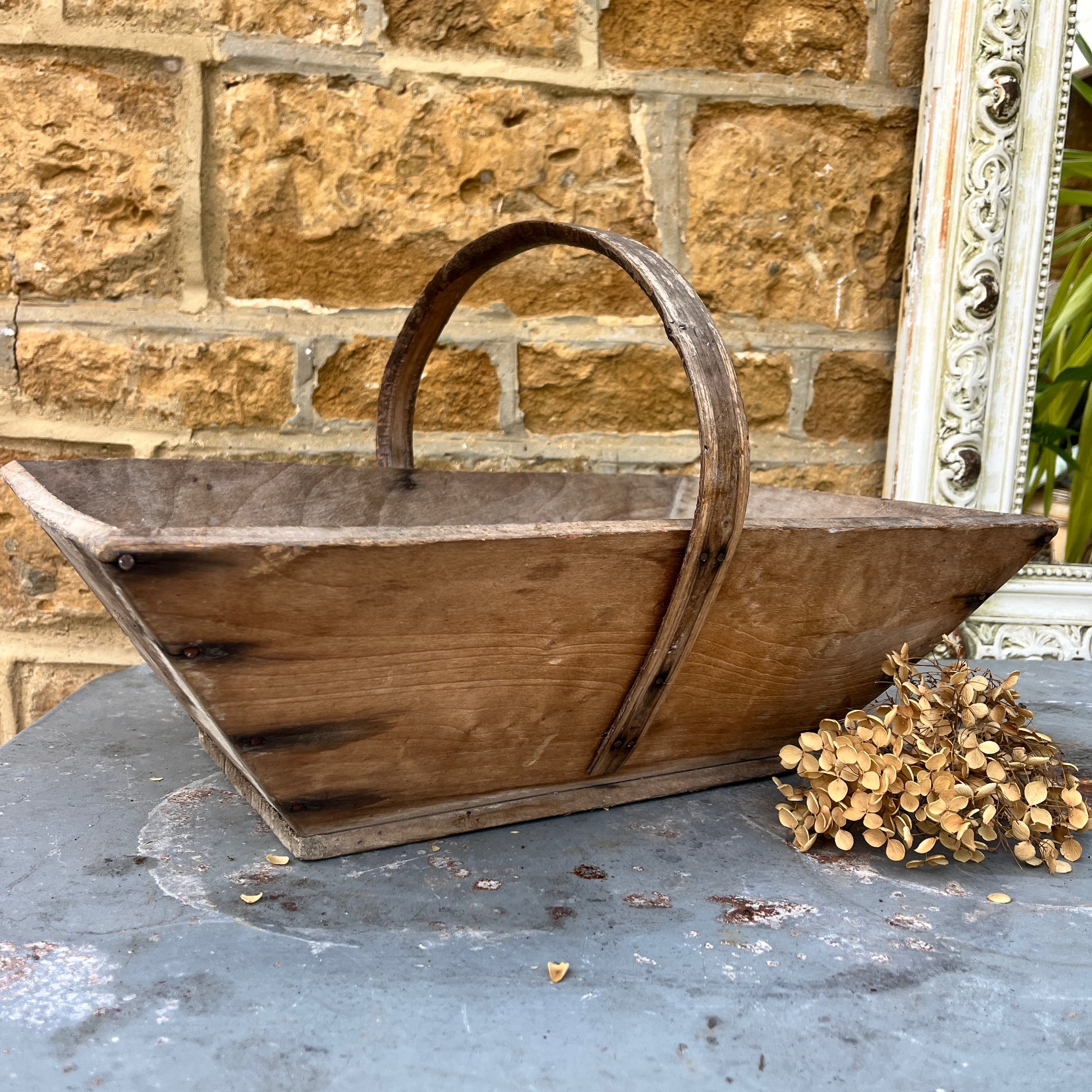 A vintage wooden French trug, featuring a shallow, rectangular shape with a sturdy central handle. The trug has a weathered, natural finish,  exuding classic gardening charm