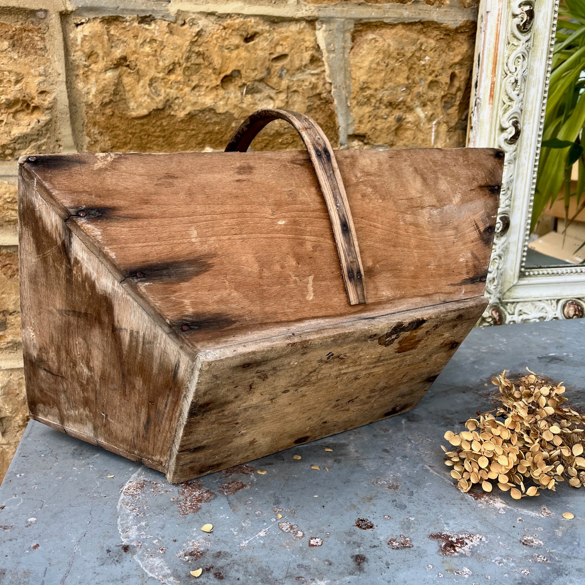 A vintage wooden French trug, featuring a shallow, rectangular shape with a sturdy central handle. The trug has a weathered, natural finish,  exuding classic gardening charm