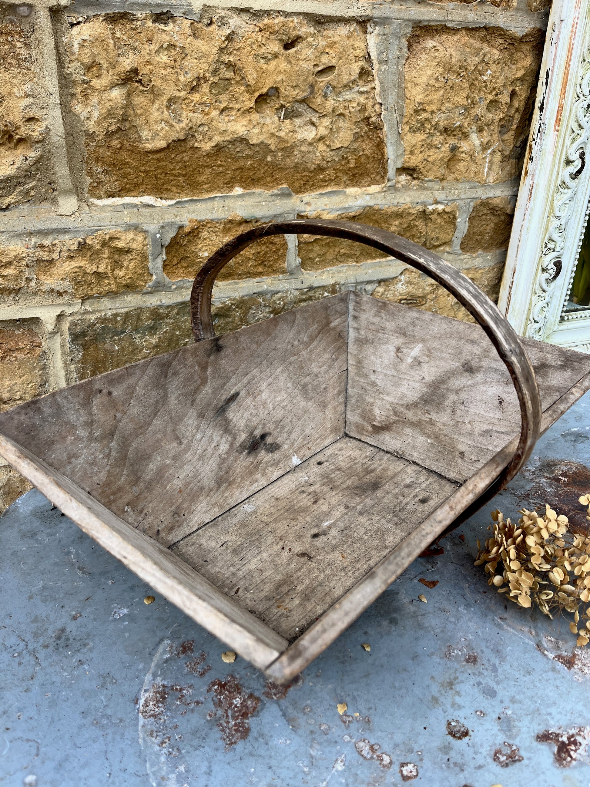 A vintage wooden French trug, featuring a shallow, rectangular shape with a sturdy central handle. The trug has a weathered, natural finish,  exuding classic gardening charm