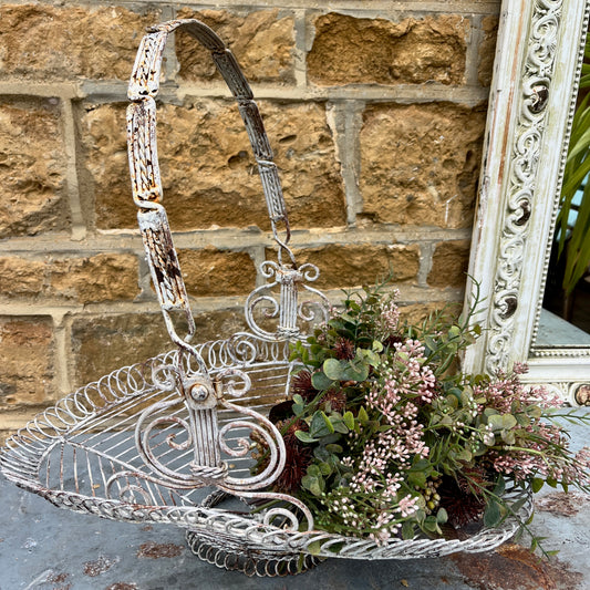 A French wire basket with a white, weathered finish, featuring an open, lattice-like design with delicate, curved edges. The basket includes one sturdy hinged handle, exuding rustic charm