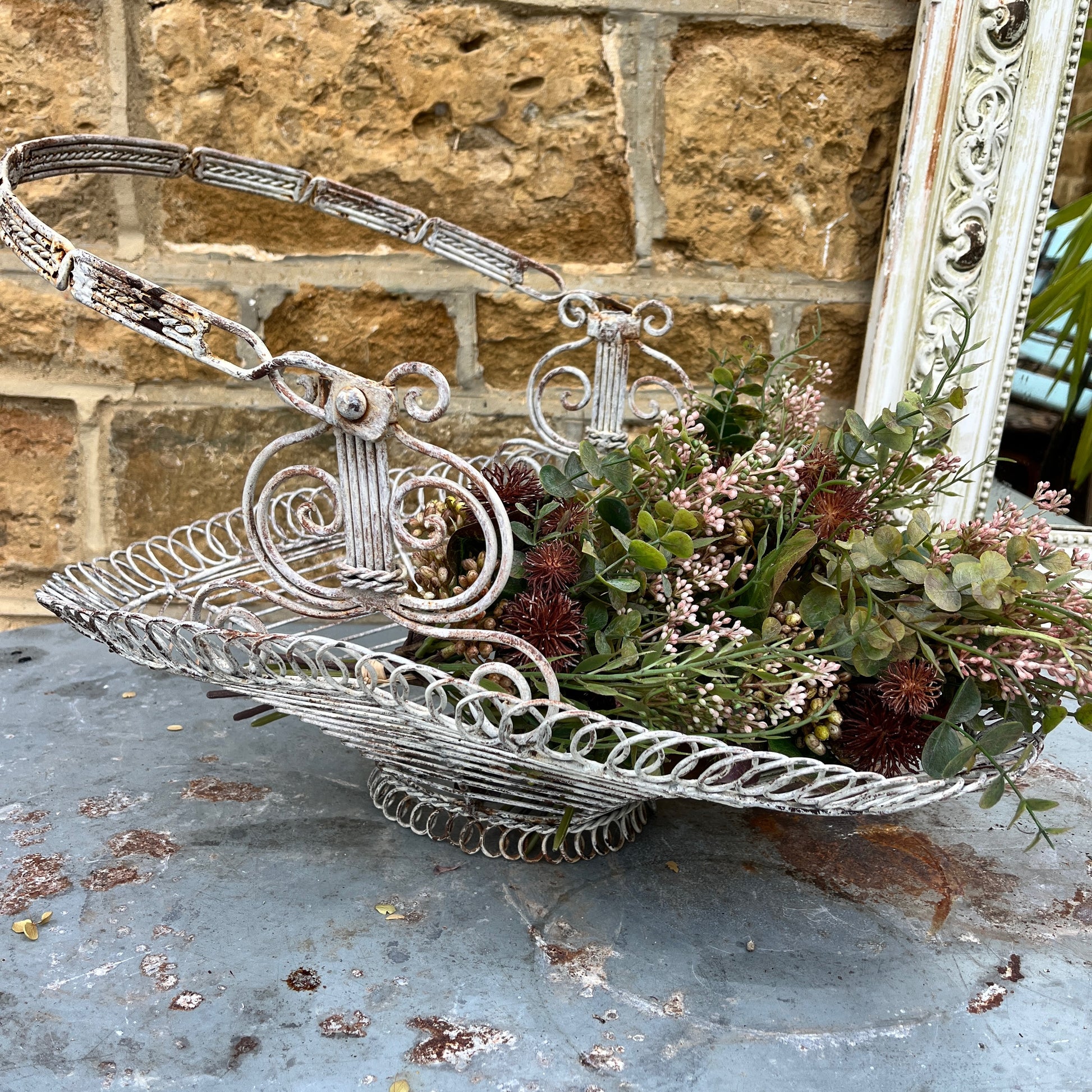 A French wire basket with a white, weathered finish, featuring an open, lattice-like design with delicate, curved edges. The basket includes one sturdy hinged handle, exuding rustic charm