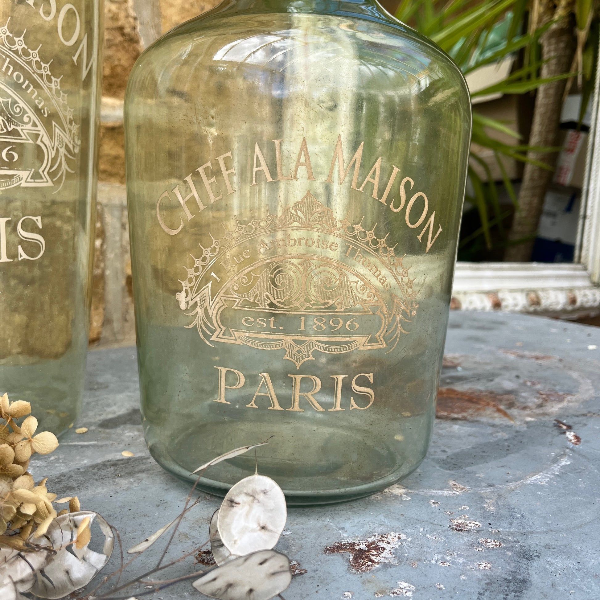 A large light green glass bottle with a smooth, glossy finish, featuring elegant gold stencil detailing of French writing and geometric patterns around its midsection.