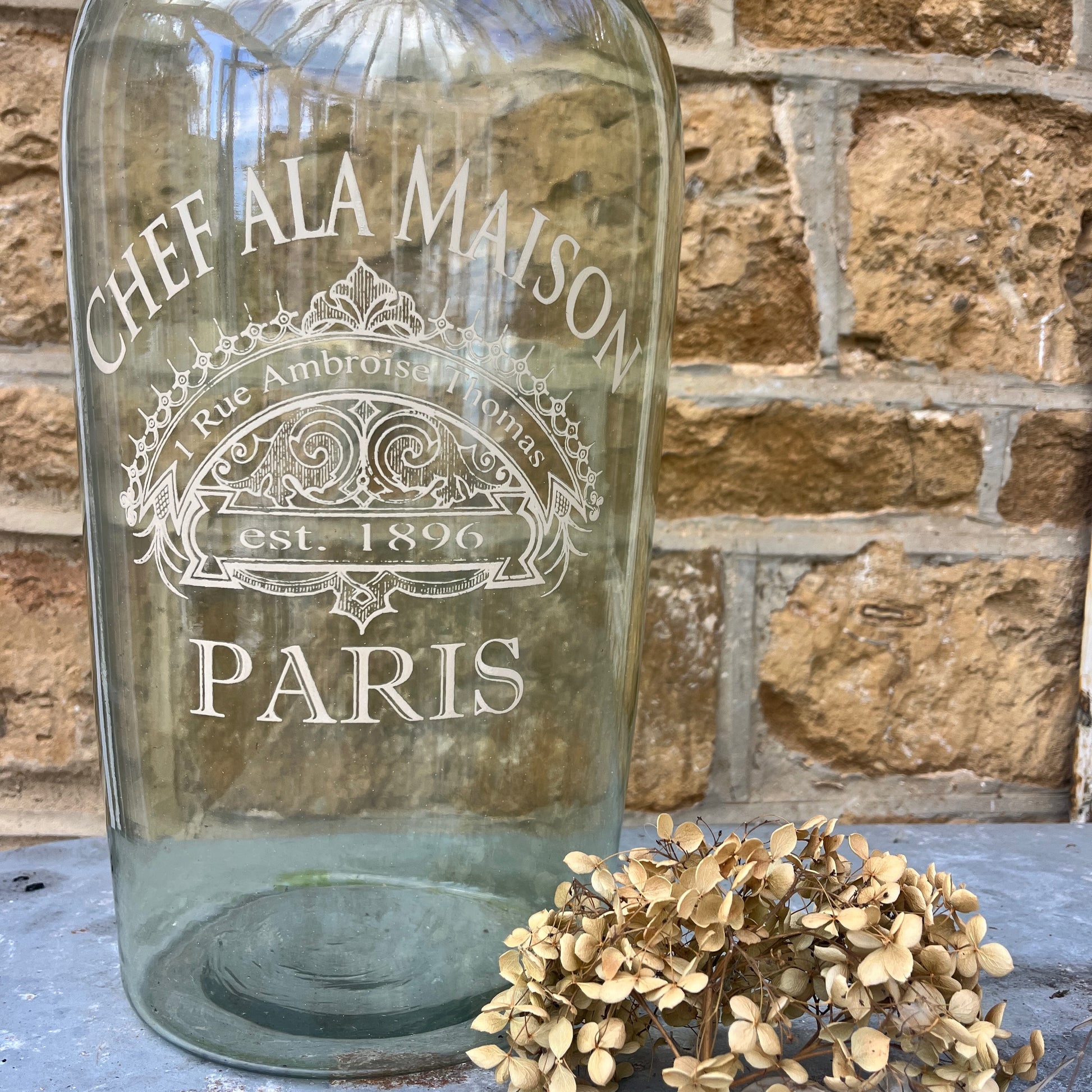 A large light green glass bottle with a smooth, glossy finish, featuring elegant gold stencil detailing of French writing and geometric patterns around its midsection.