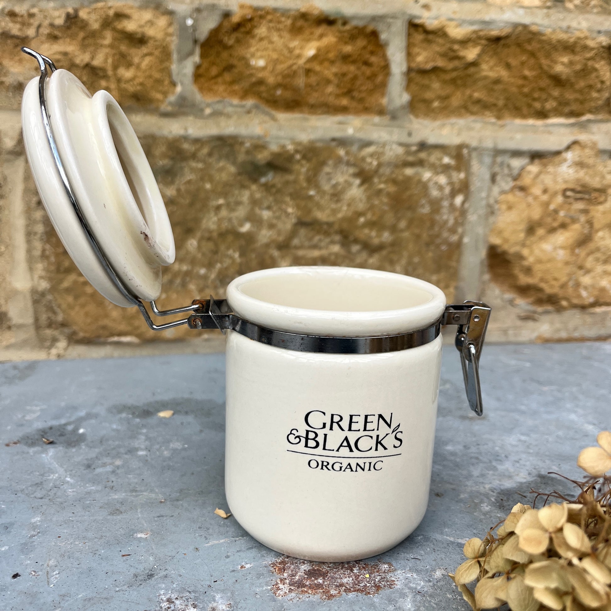 A ceramic jar,  with a hinged lid featuring a classic design, labeled Green & Black's organic. The jar has a vintage, sturdy look.