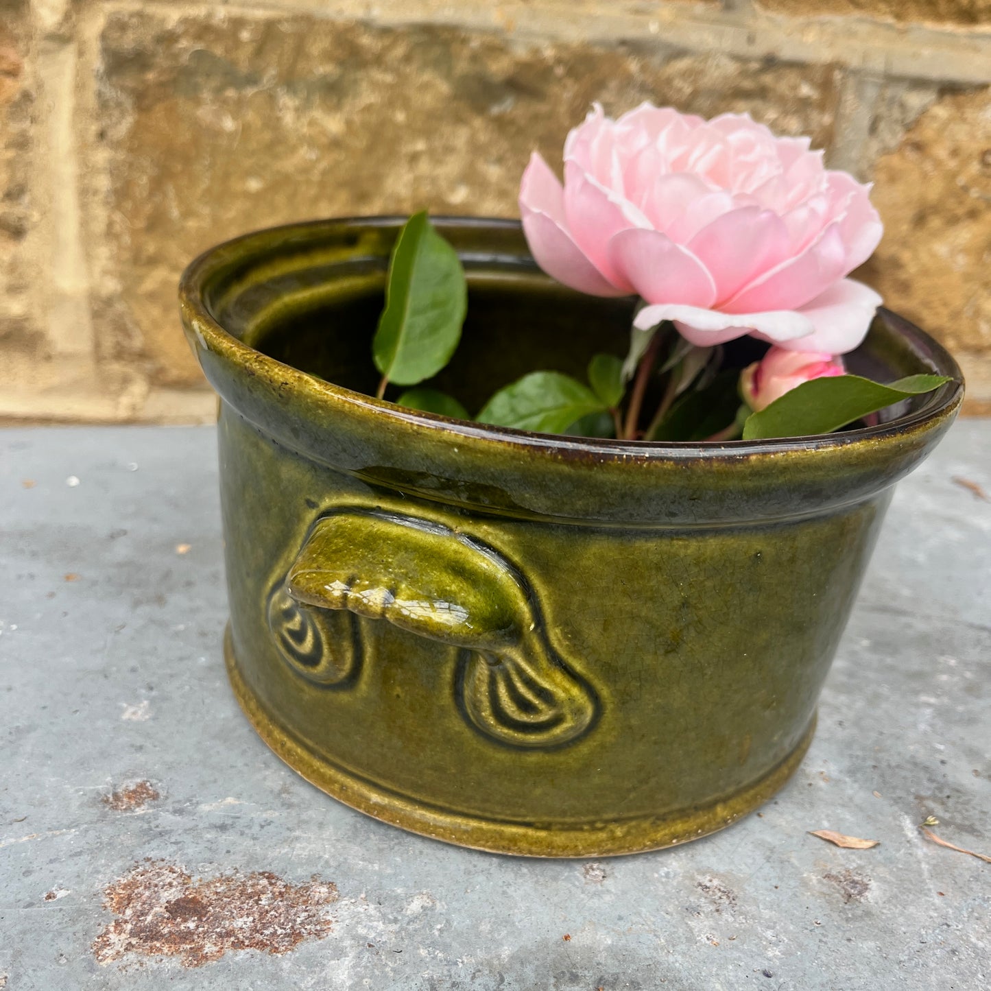 Green ceramic planter with a pink flower against a stone wall background