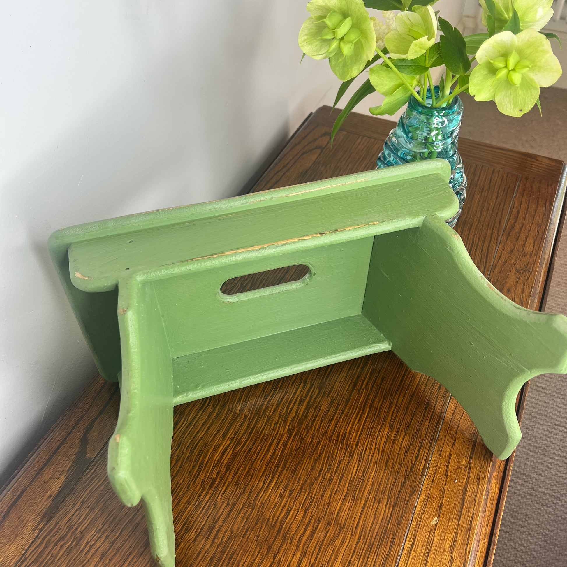 A rustic wooden stool painted in a weathered green finish, showcasing a distressed texture with visible wood grain.