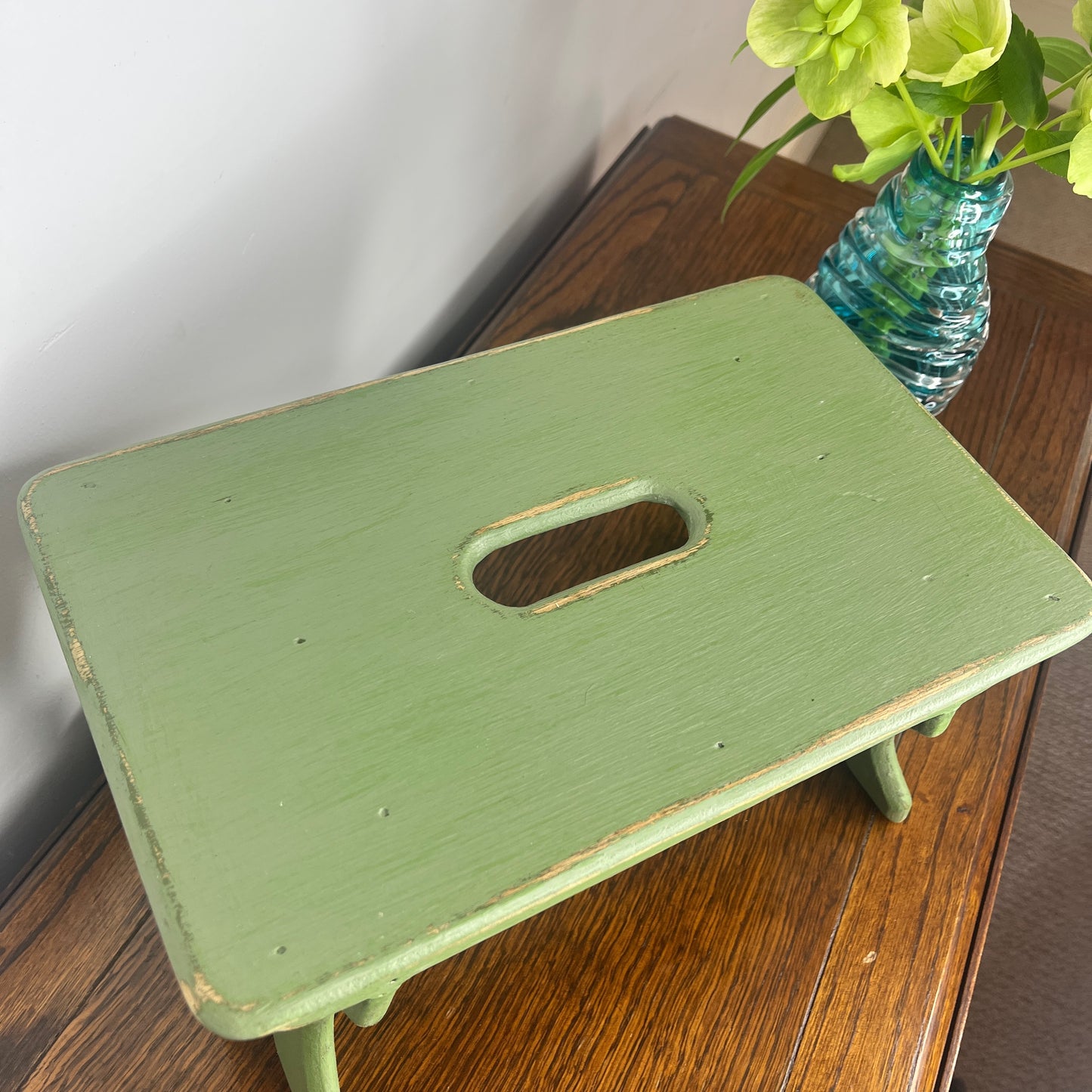 A rustic wooden stool painted in a weathered green finish, showcasing a distressed texture with visible wood grain.