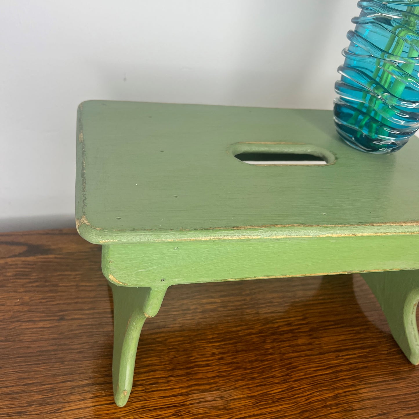 A rustic wooden stool painted in a weathered green finish, showcasing a distressed texture with visible wood grain.