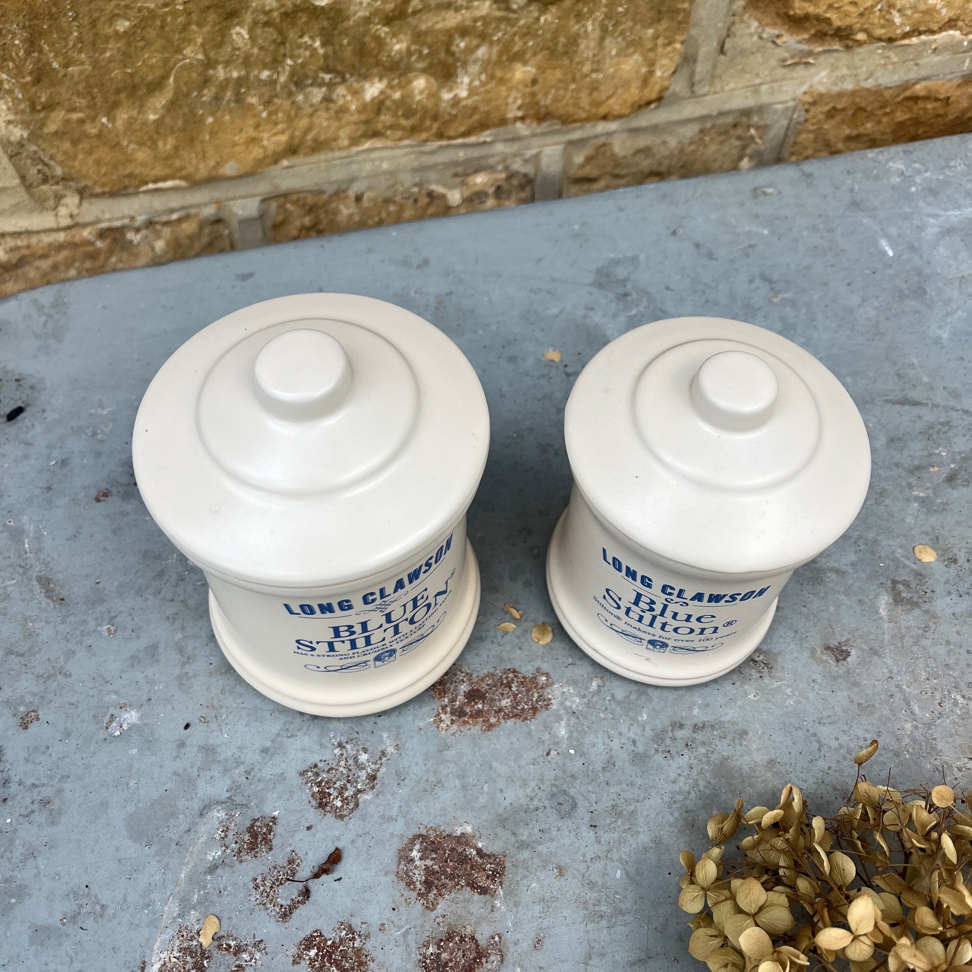 A pair of ceramic jars, each with a fitted lid featuring a classic design, labeled Blue Stilton cheese. The jars have a vintage, sturdy look.