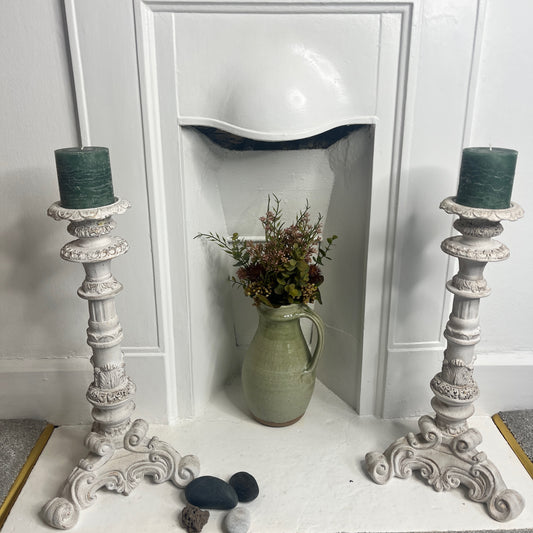 A pair of tall French-style candle holders crafted from wood, painted white, featuring elegant, carved detailing and a slightly distressed finish. Each holder supports a single candle, exuding classic rustic charm