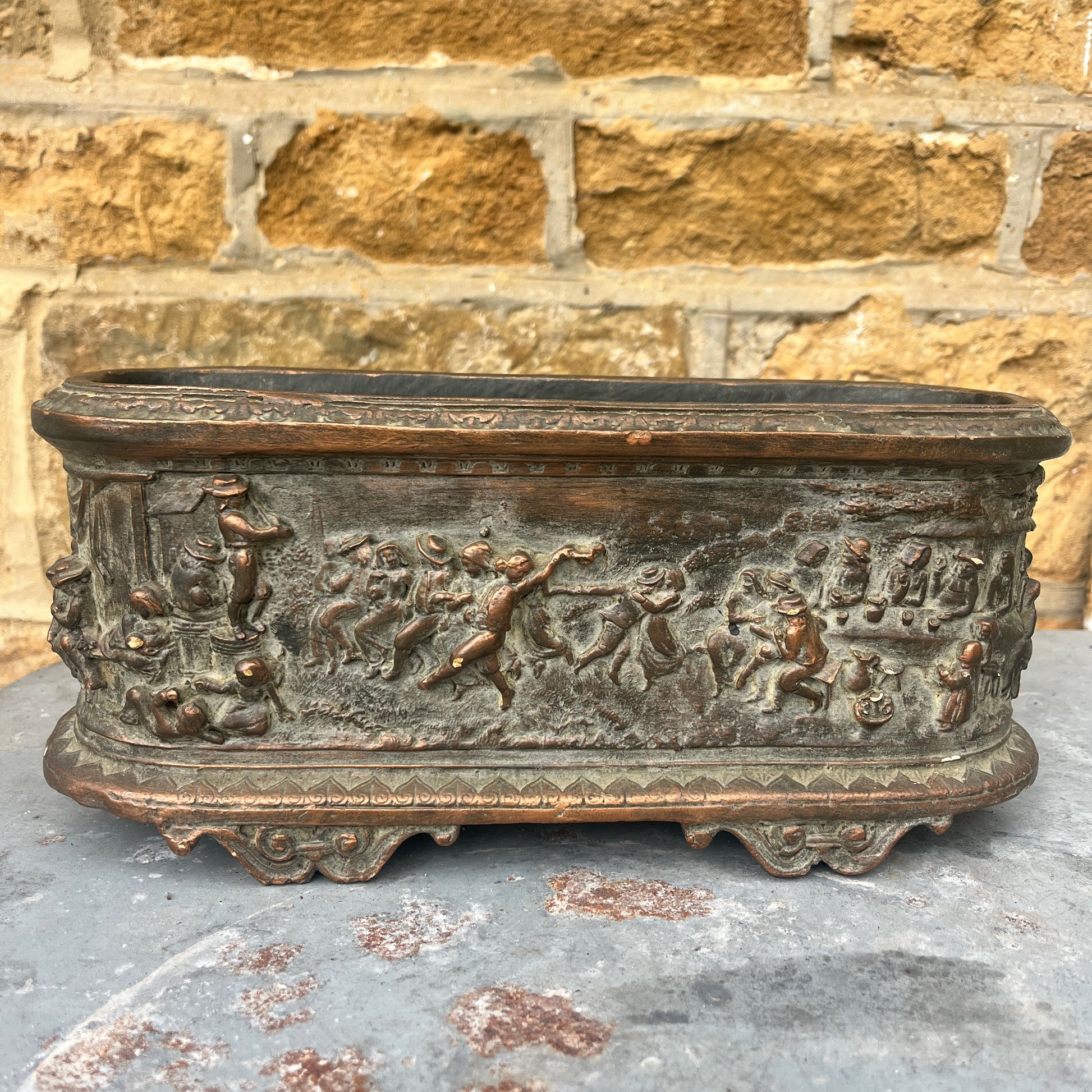 A vintage terracotta jardinière with a rich, earthy finish, featuring an intricately moulded frieze scene of classical figures and animals encircling its exterior. The cylindrical planter has a slightly weathered texture, exuding rustic elegance