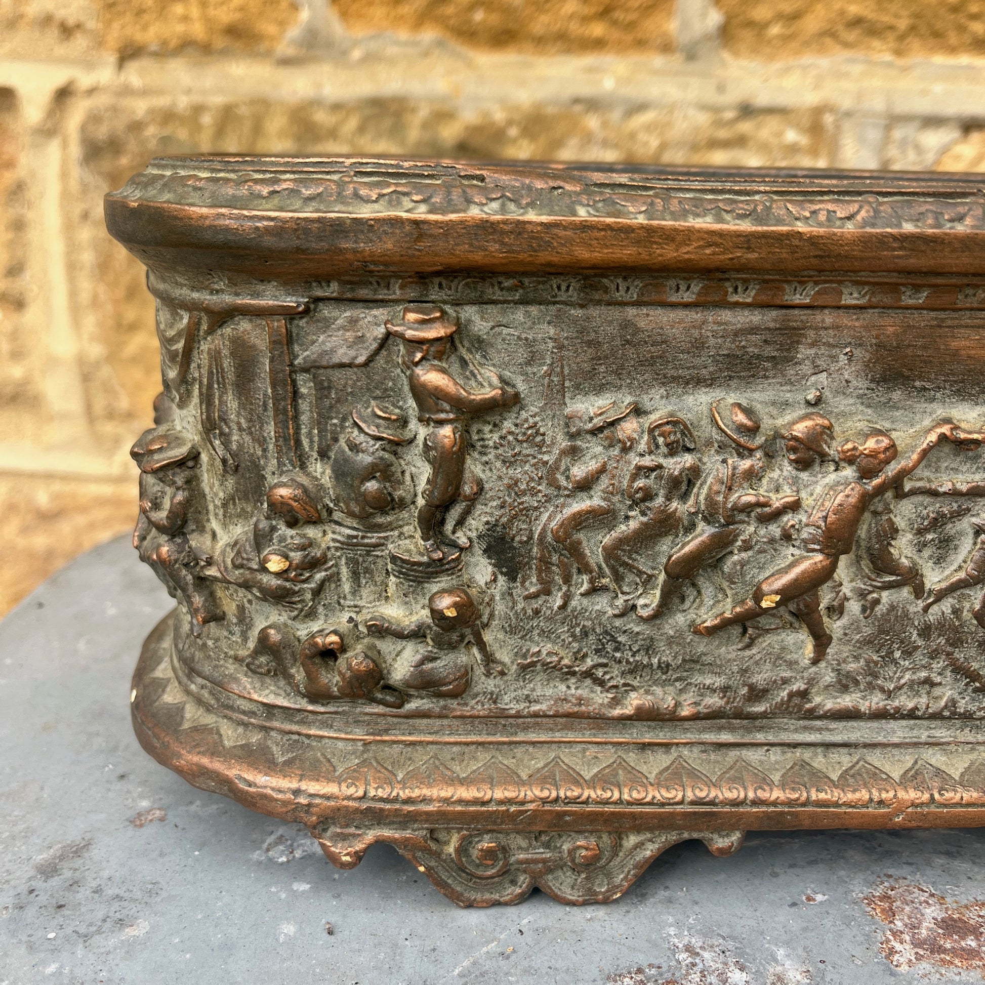 A vintage terracotta jardinière with a rich, earthy finish, featuring an intricately moulded frieze scene of classical figures and animals encircling its exterior. The cylindrical planter has a slightly weathered texture, exuding rustic elegance