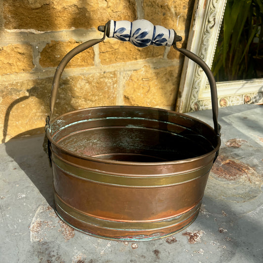 A vintage French copper planter with a glossy finish, featuring a delicate china handle adorned with a blue and white floral pattern.