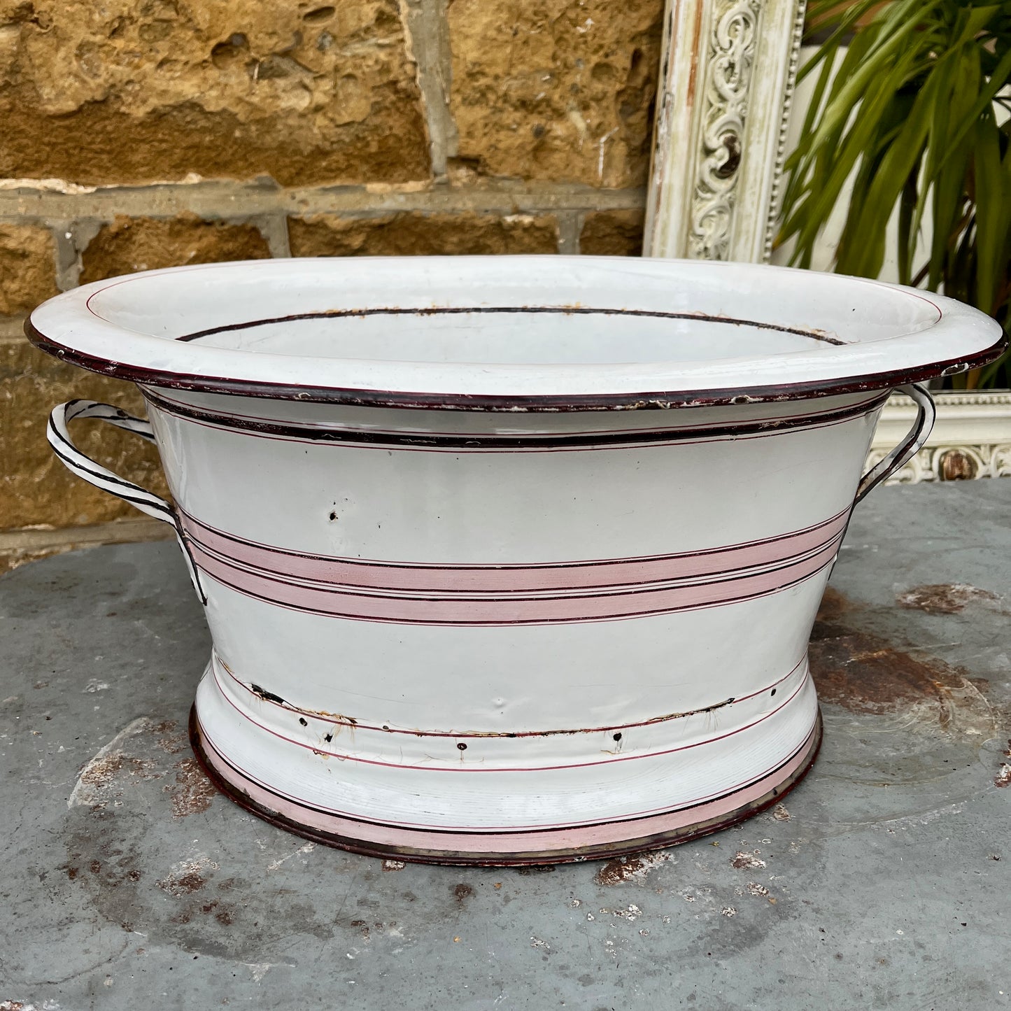 A large vintage French white enamel wash bowl, circa early 20th century, featuring a wide, deep basin. The bowl is adorned with delicate pink stripes,  with a smooth white enamel finish showing minor chips and rust spots, characteristic of its aged, rustic charm.