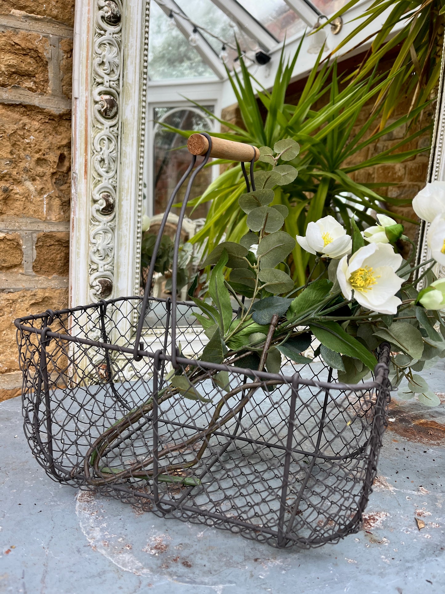 A rustic wire basket with a sturdy, woven metal frame, featuring a natural wooden handle across the top. The open, grid-like design of the basket reveals its contents, exuding a simple, farmhouse charm.