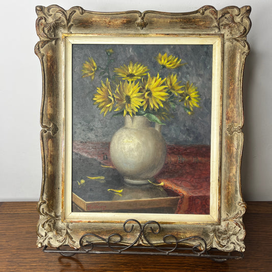 A vintage still life oil on board painting depicting a delicate bouquet of blooming yellow chrysanthemums in a grey vase, set against a softly lit table with subtle shadows and aged patina.