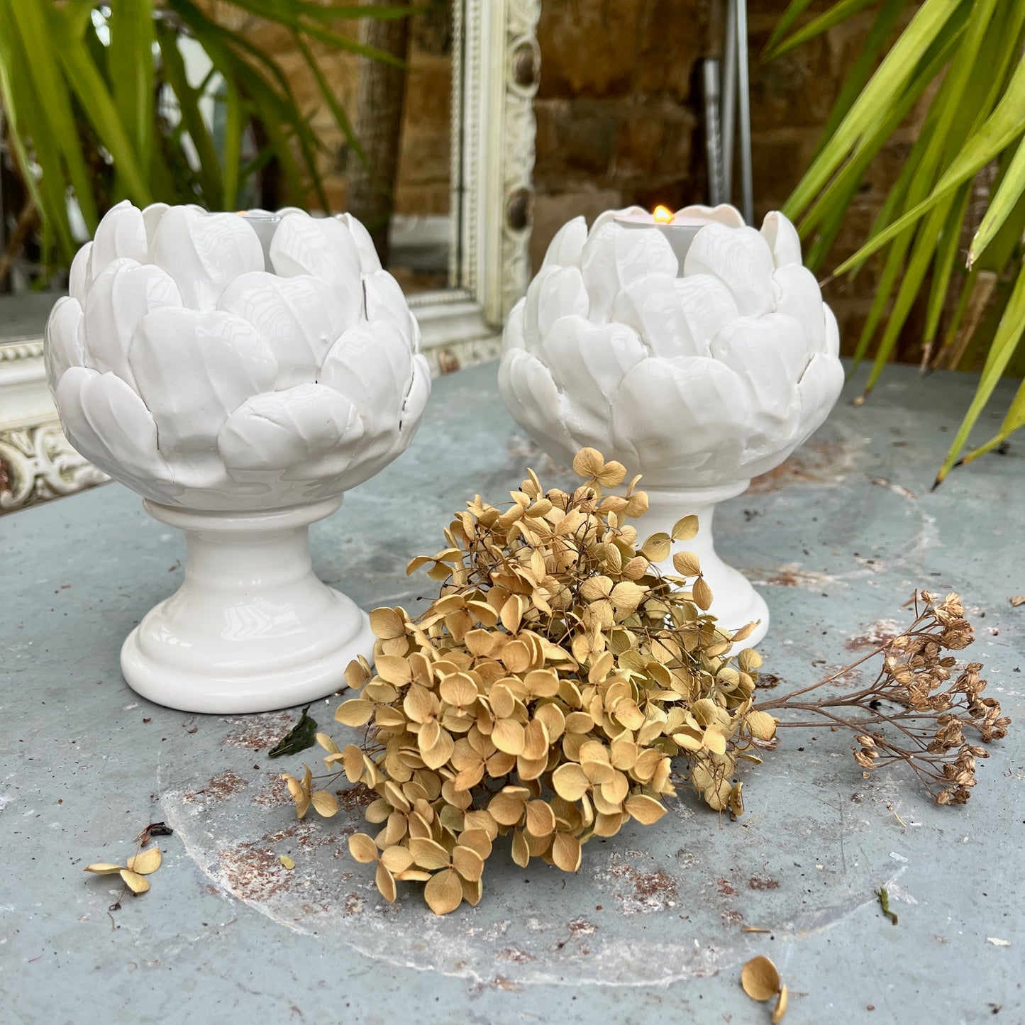 A ceramic candle holder sculpted in the shape of an artichoke, featuring detailed, layered leaves in a soft white glaze. The holder cradles a single tealight candle at its center, exuding a natural, organic charm
