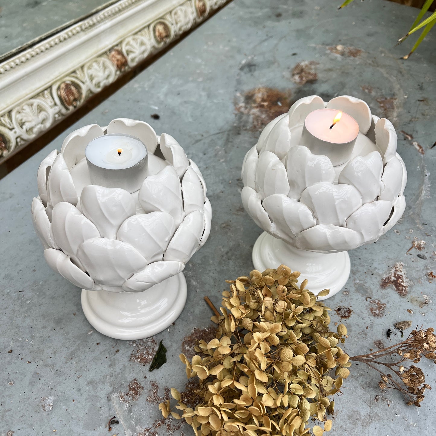 A ceramic candle holder sculpted in the shape of an artichoke, featuring detailed, layered leaves in a soft white glaze. The holder cradles a single tealight candle at its center, exuding a natural, organic charm