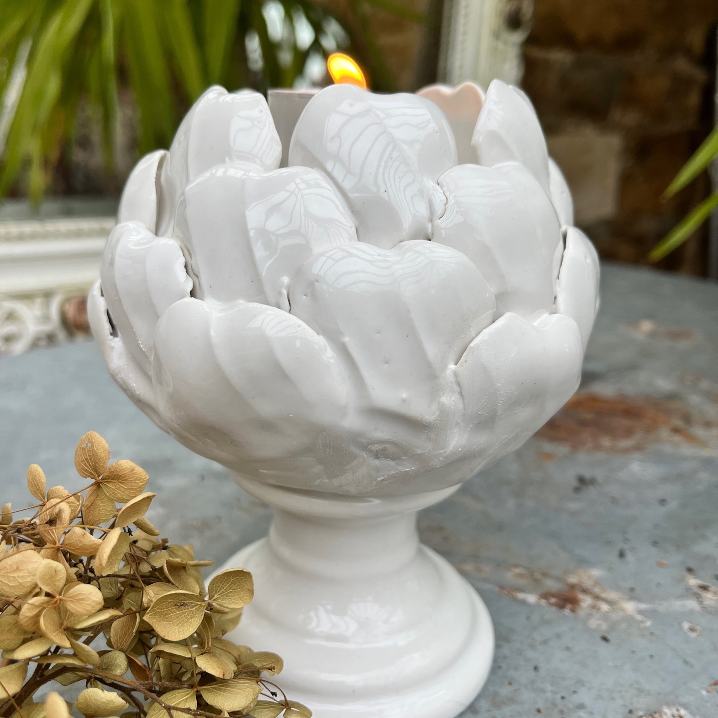 A ceramic candle holder sculpted in the shape of an artichoke, featuring detailed, layered leaves in a soft white glaze. The holder cradles a single tealight candle at its center, exuding a natural, organic charm