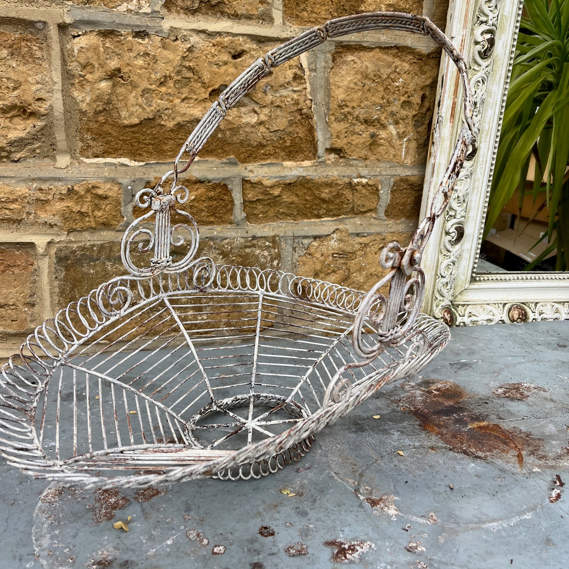 A French wire basket with a white, weathered finish, featuring an open, lattice-like design with delicate, curved edges. The basket includes one sturdy hinged handle, exuding rustic charm