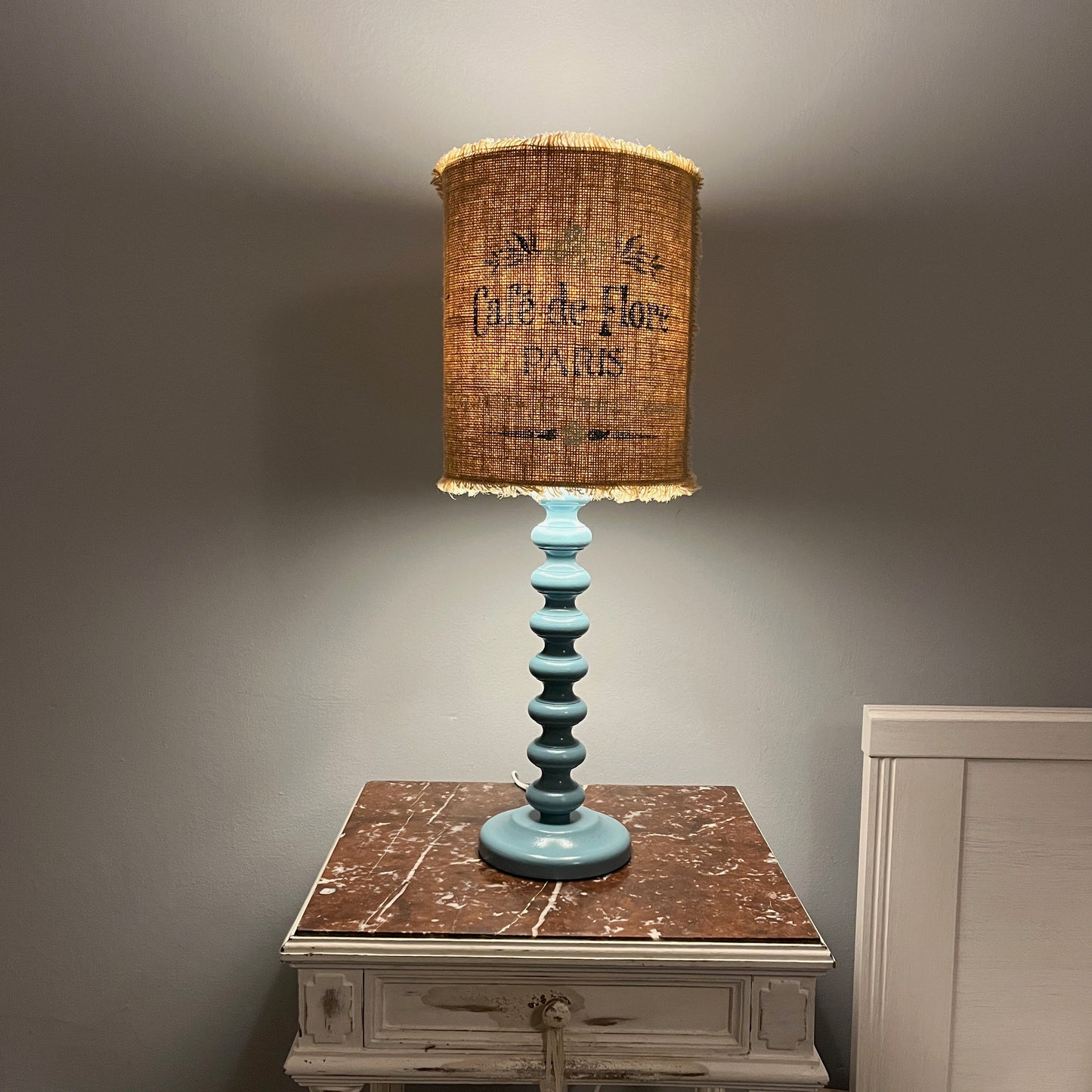 A handmade lampshade crafted from hessian fabric which has been stencilled, which offers a rustic texture and warm tone, paired with a hand-painted bobble base for a cozy, artisanal look.