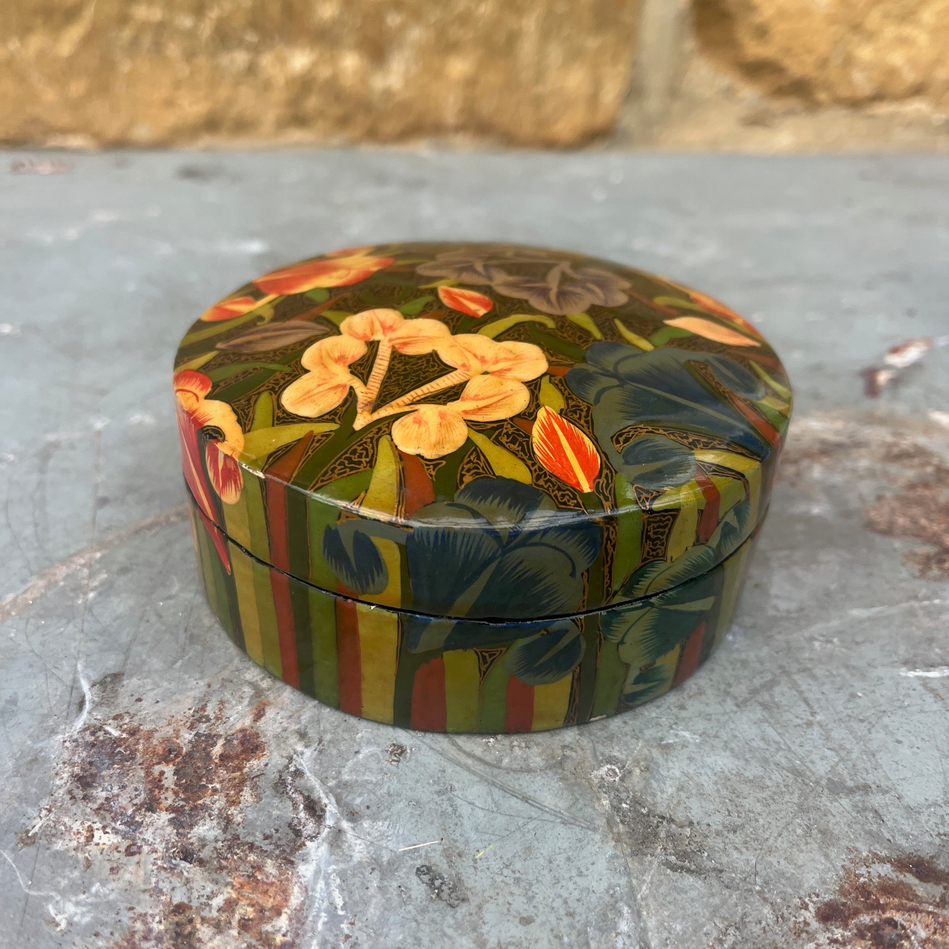 A set of round papier-mâché coasters, hand-painted with colorful floral designs in shades of yellow, red, blue and green, neatly stacked inside a matching rectangular papier-mâché box.