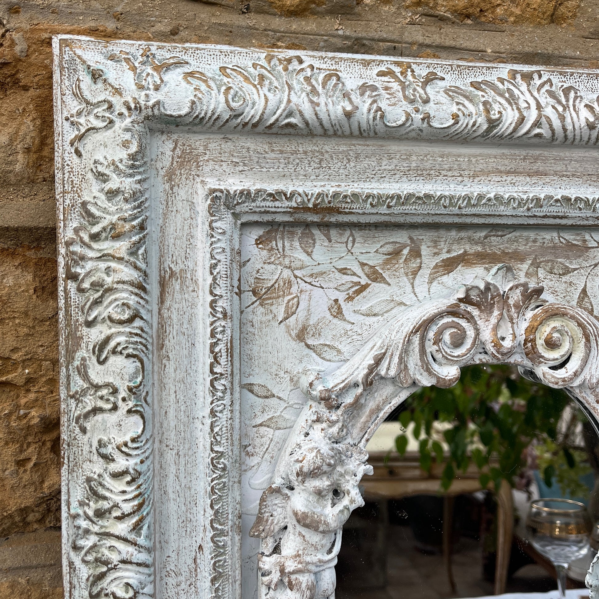 A shabby chic rectangular mirror with a distressed, white wooden frame, adorned with small, intricately carved angel figures positioned around the edge of the mirror. The vintage-style mirror reflects a soft, weathered charm.