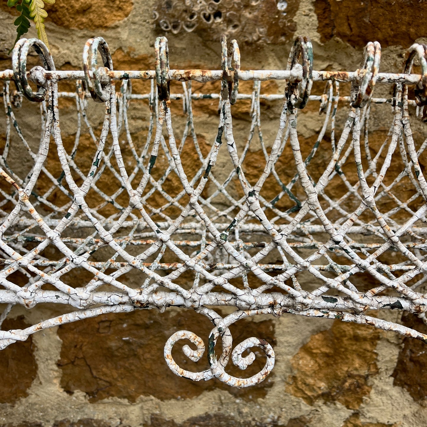 A Victorian decorative plant stand crafted from ornate wrought iron wire, featuring intricate scrollwork and floral patterns. The two-tiered stand, painted in a distressed white finish, supports small potted plants, exuding elegant vintage charm